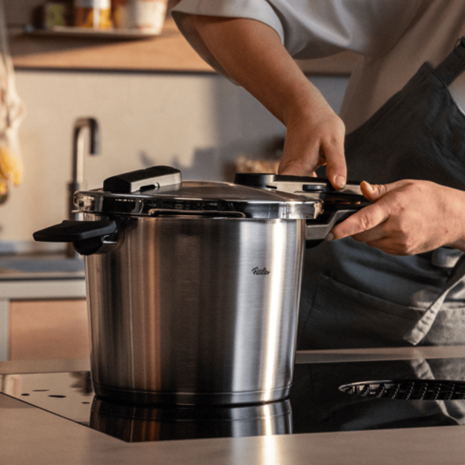 Eine Person in einer grauen Schürze befestigt den Deckel eines Fissler Schnellkochtopfs aus Edelstahl auf einer Herdplatte in einer modernen Küche.
