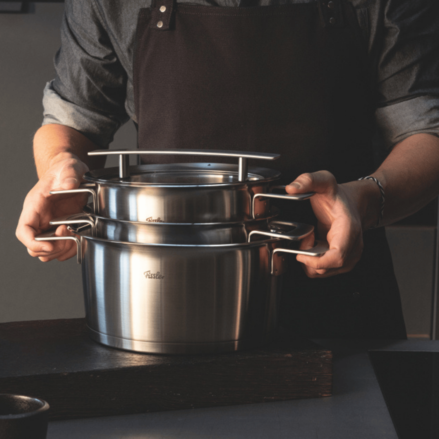 Eine Person in einer dunklen Schürze hält zwei übereinander gestapelte Fissler-Edelstahltöpfe mit Deckeln, deren polierte Oberflächen im sanften Licht schimmern.