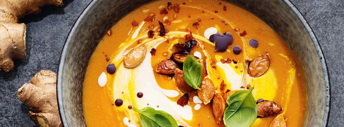 Eine Schüssel mit einer reichhaltigen, cremigen Orangensuppe, elegant angerichtet mit Basilikumblättern, Kernen, zarten Sahnespiralen und einem Hauch Öl, dazu frische Ingwerstückchen auf einem dunklen Untergrund. Zubereitet und serviert mit dem präzisionsgefertigten Kochgeschirr von Fissler für optimalen Geschmack und zeitlosen Stil.