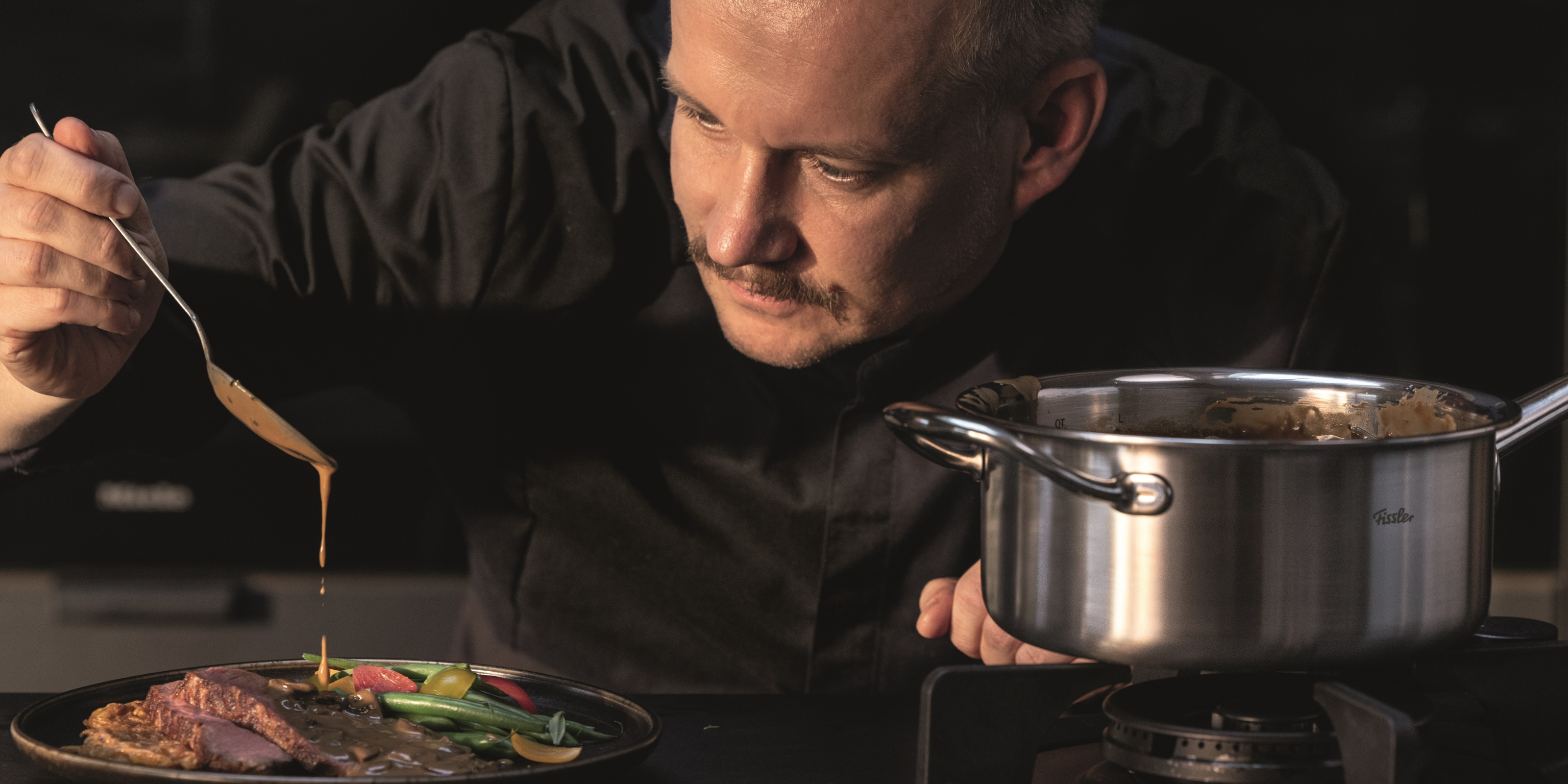 Ein Koch serviert seine Speisen präzise mit einem Löffel, während ein Fissler-Edelstahltopf auf dem Herd in einer modernen Küche steht.