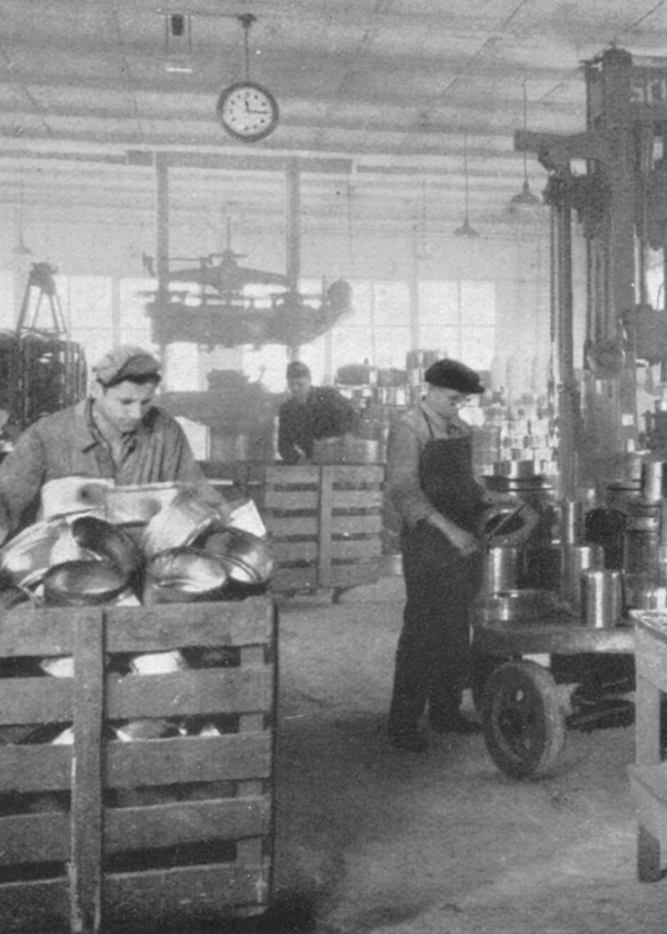 Historisches Schwarz-Weiß-Foto von Fissler-Handwerkern in einer Fabrik, die inmitten alter Maschinen fachmännisch Kochgeschirr aus Edelstahl zusammenbauen.