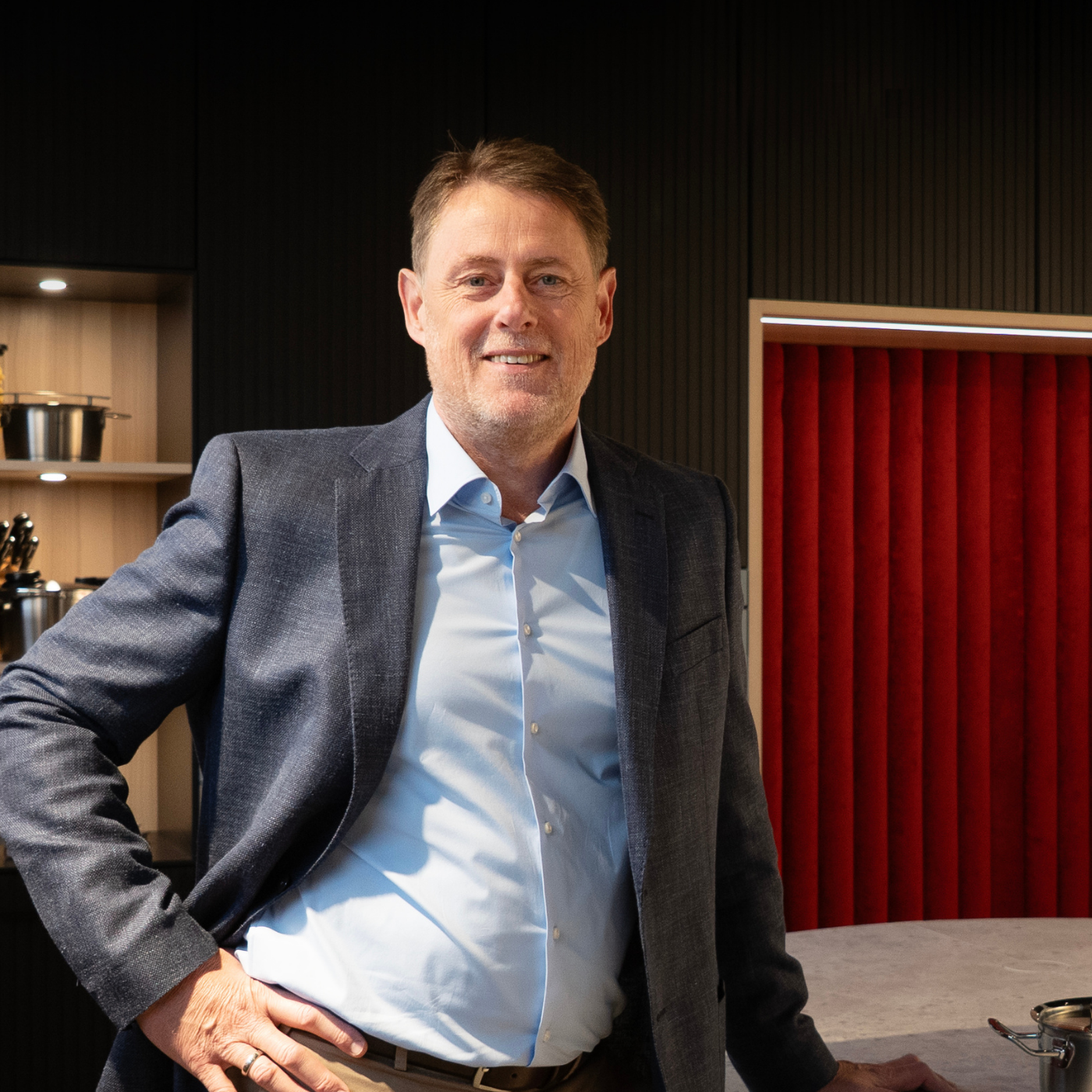 Ein Mann in einem blauen Blazer steht lächelnd an einer Küchentheke mit Fissler-Edelstahltöpfen und dem charakteristischen roten Branding im Hintergrund.
