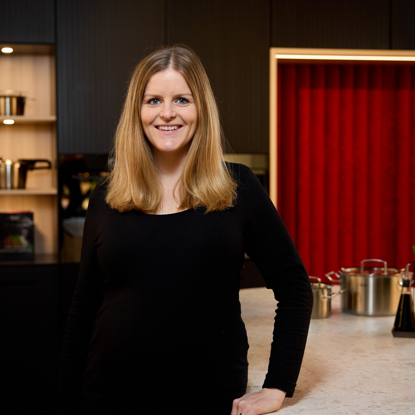 Eine Frau lächelt in einer modernen Küche, in der Fissler-Edelstahltöpfe auf dem Tresen inmitten schlichter dunkler Schränke und roter Akzente stehen.
