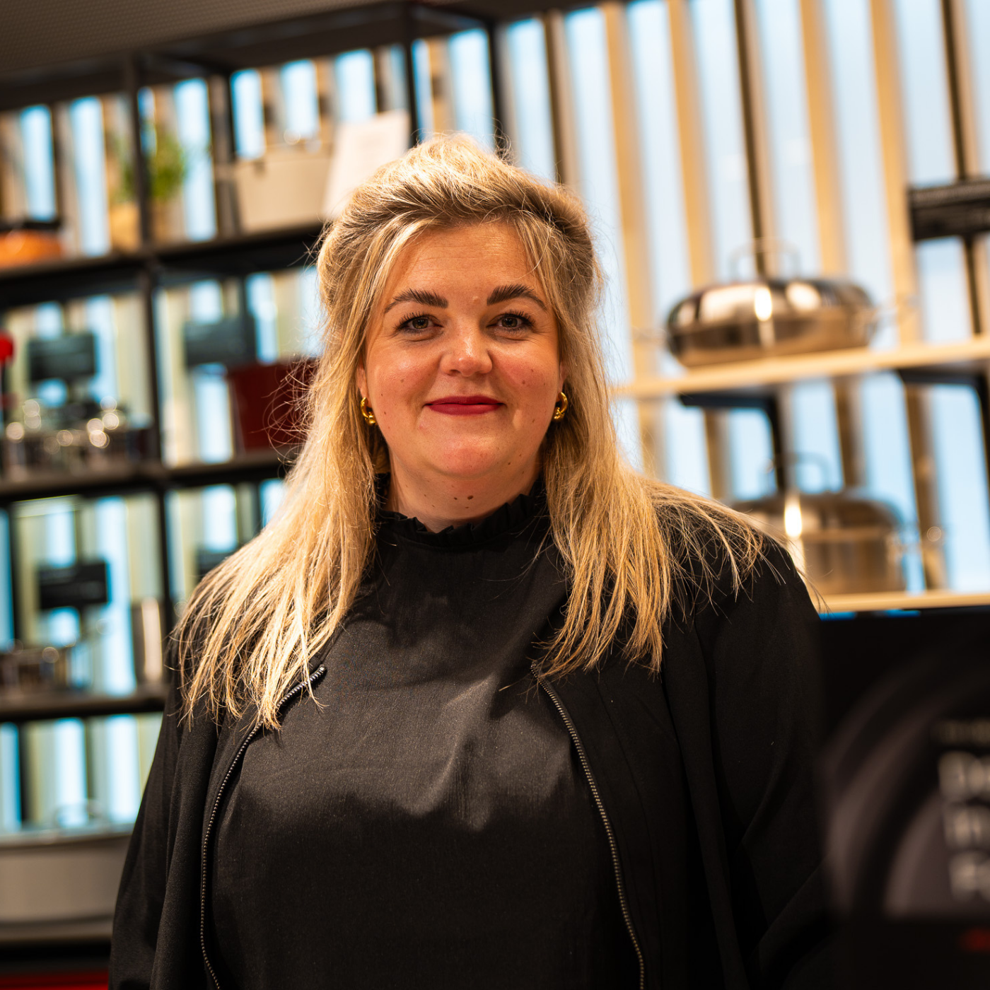 Eine Frau lächelt in einer hellen, modernen Küche, hinter der in Regalen Töpfe und Pfannen aus rostfreiem Stahl von Fissler stehen.