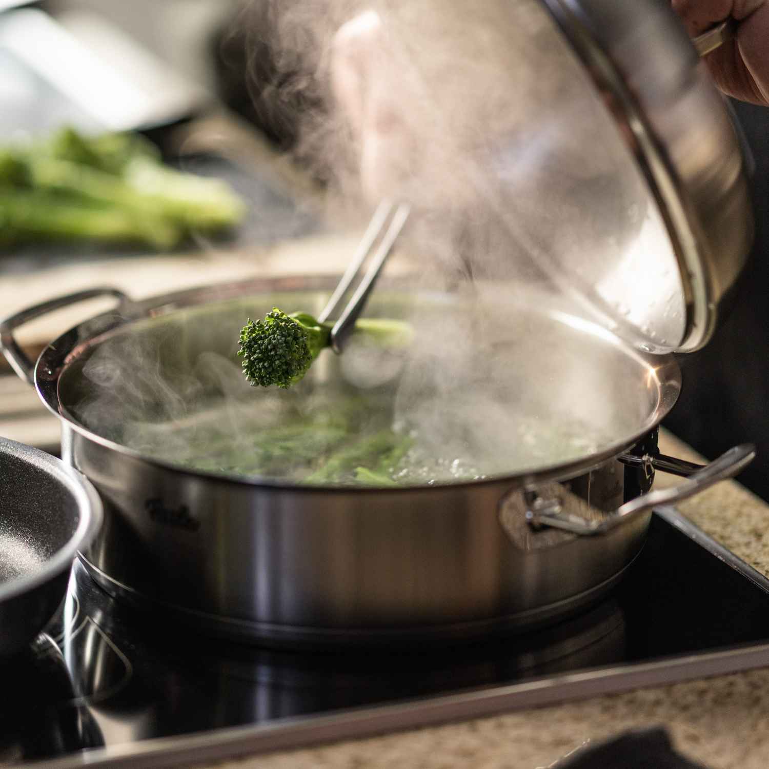A chef opens a steaming Fissler stainless steel pot on the stove, holding broccoli with tongs. Fresh vegetables highlight the quality of the kitchen.