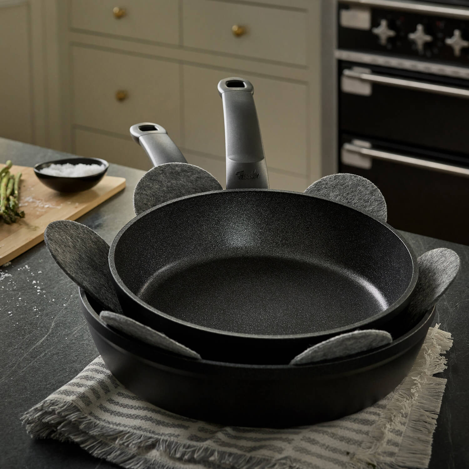 Two Fissler non-stick pans with a gray pan protector, stacked on a striped cloth, are an expression of German innovation and timeless design.