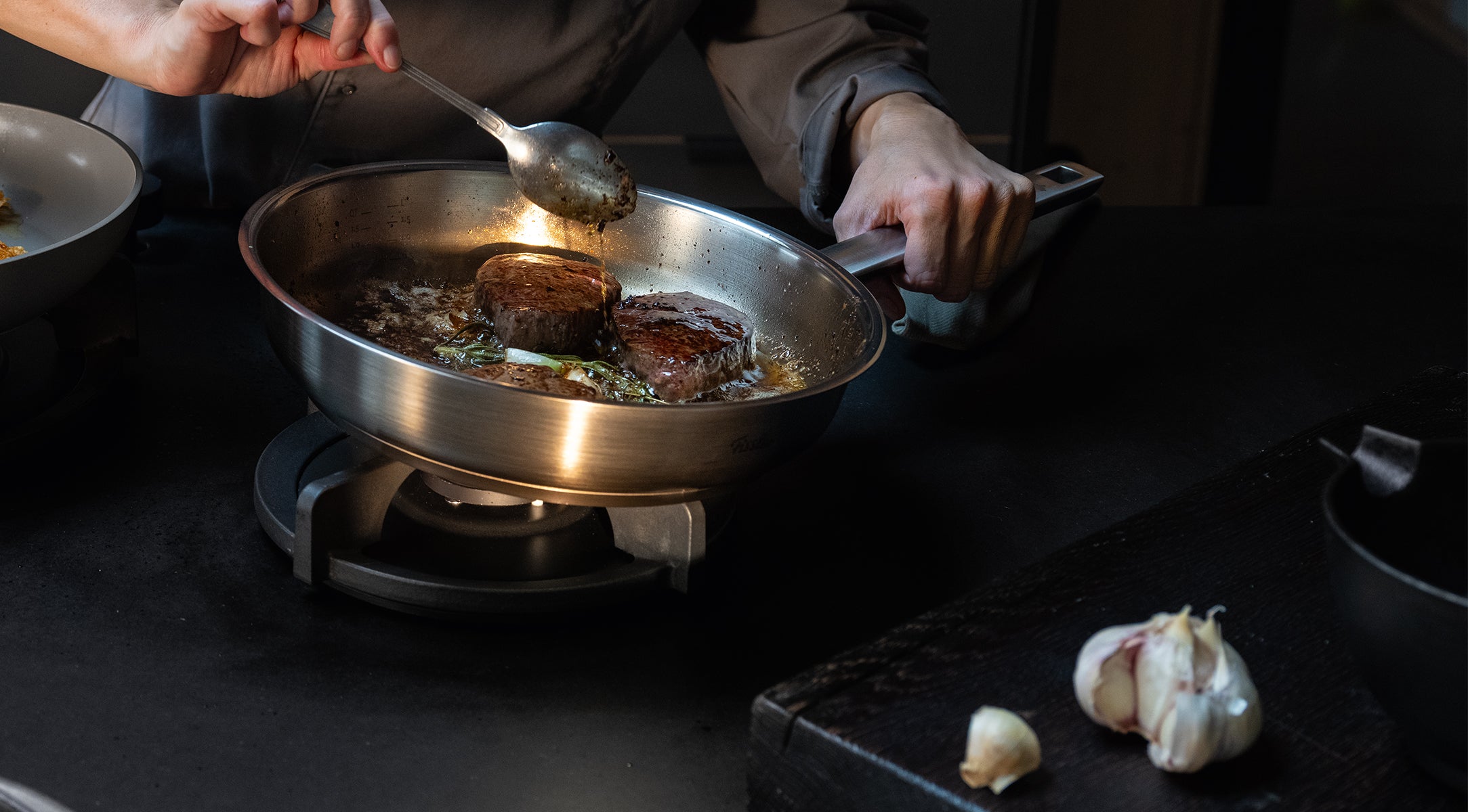 A person expertly searing two steaks in a premium Fissler stainless steel skillet, spooning butter over the meat as fresh herbs and garlic sizzle in the pan; a whole head of garlic rests on the counter, underscoring the precision and quality of Fissler cookware "Made in Germany.