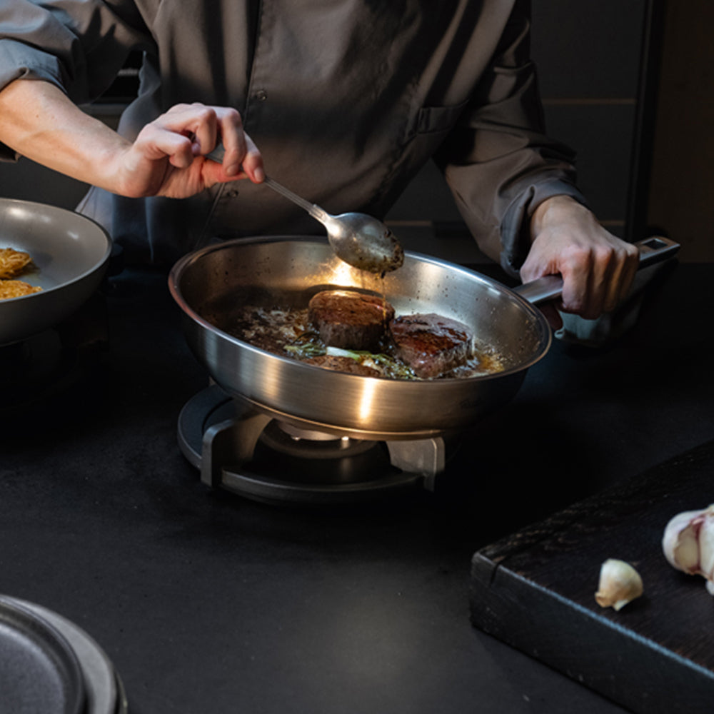 Ein Koch bereitet auf einem Herd in einer hochwertigen Fissler-Edelstahlpfanne gekonnt Steaks zu und gießt dabei sorgfältig Soße über das Fleisch. Daneben runden frischer Knoblauch und ein elegantes dunkles Schneidebrett das Bild der professionellen Küche ab und unterstreichen Fisslers Engagement für präzise Technik und zeitloses Design.