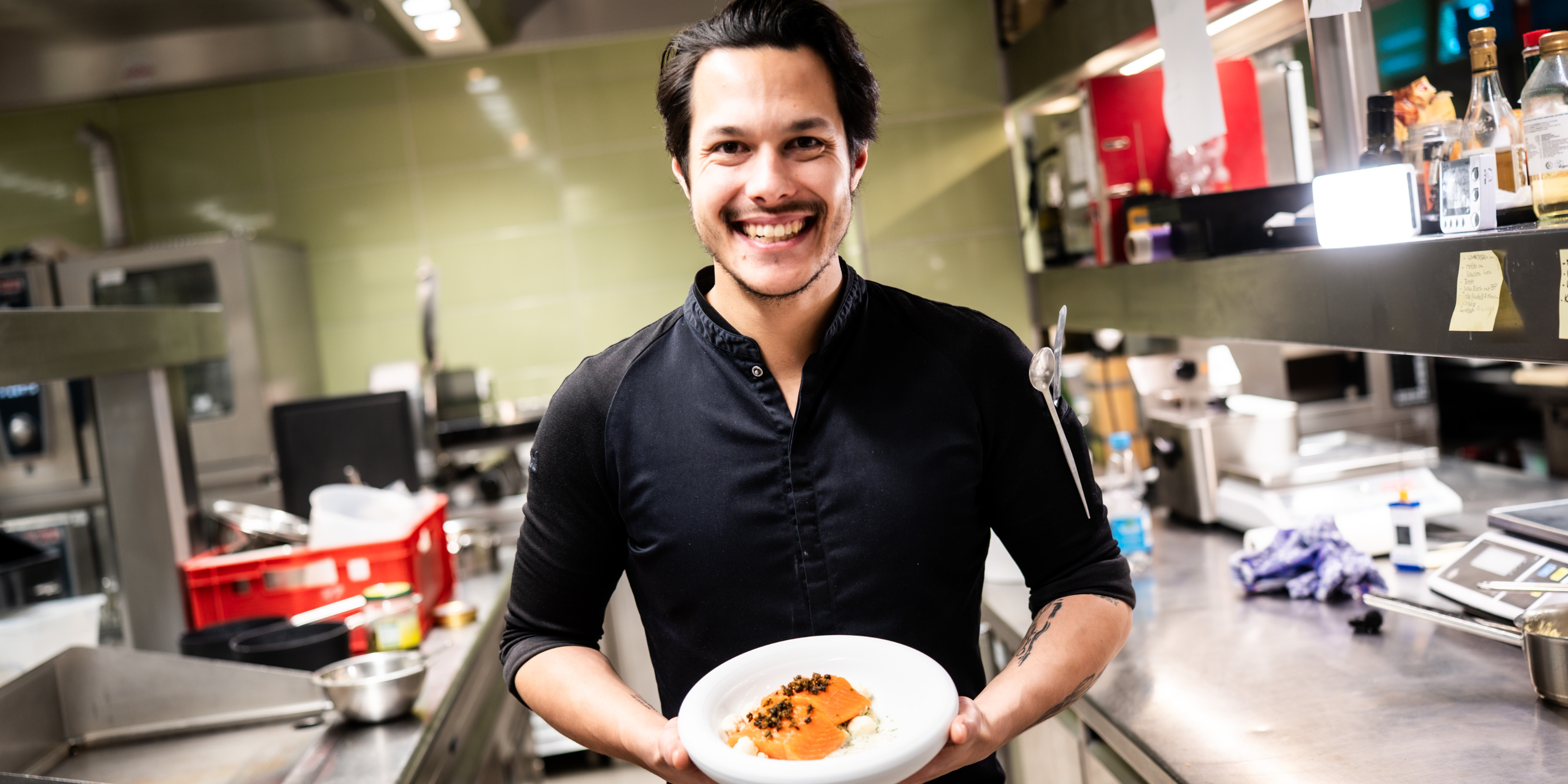 A smiling man in a black shirt holds a plate of food in a professional kitchen, surrounded by Fissler’s precision-engineered cookware, including stainless steel pots and pans, with durable utensils and innovative kitchen accessories visible in the background.