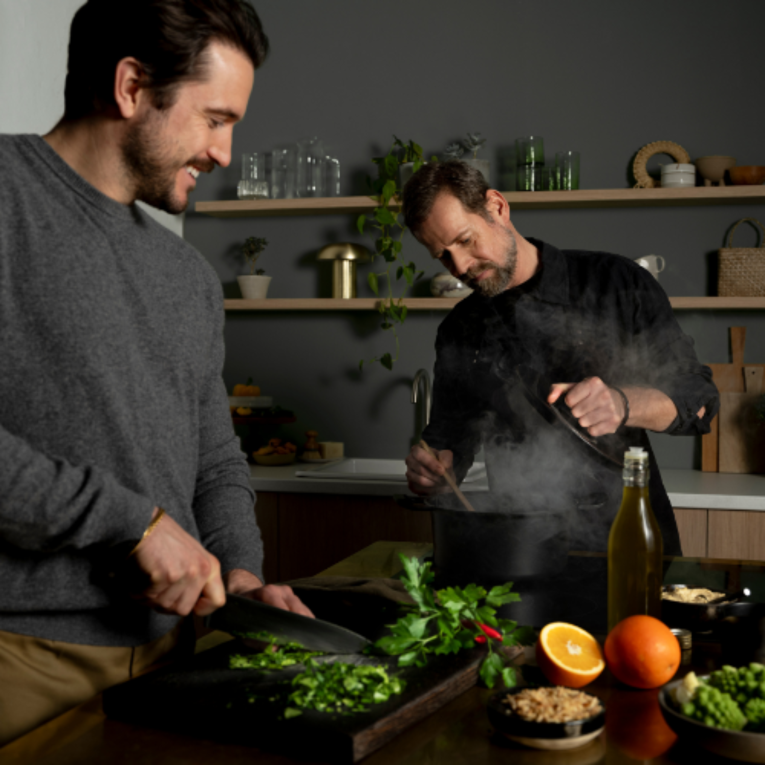 Two men cook in a modern kitchen with Fissler stainless steel pots while preparing fresh herbs and ingredients on the counter.