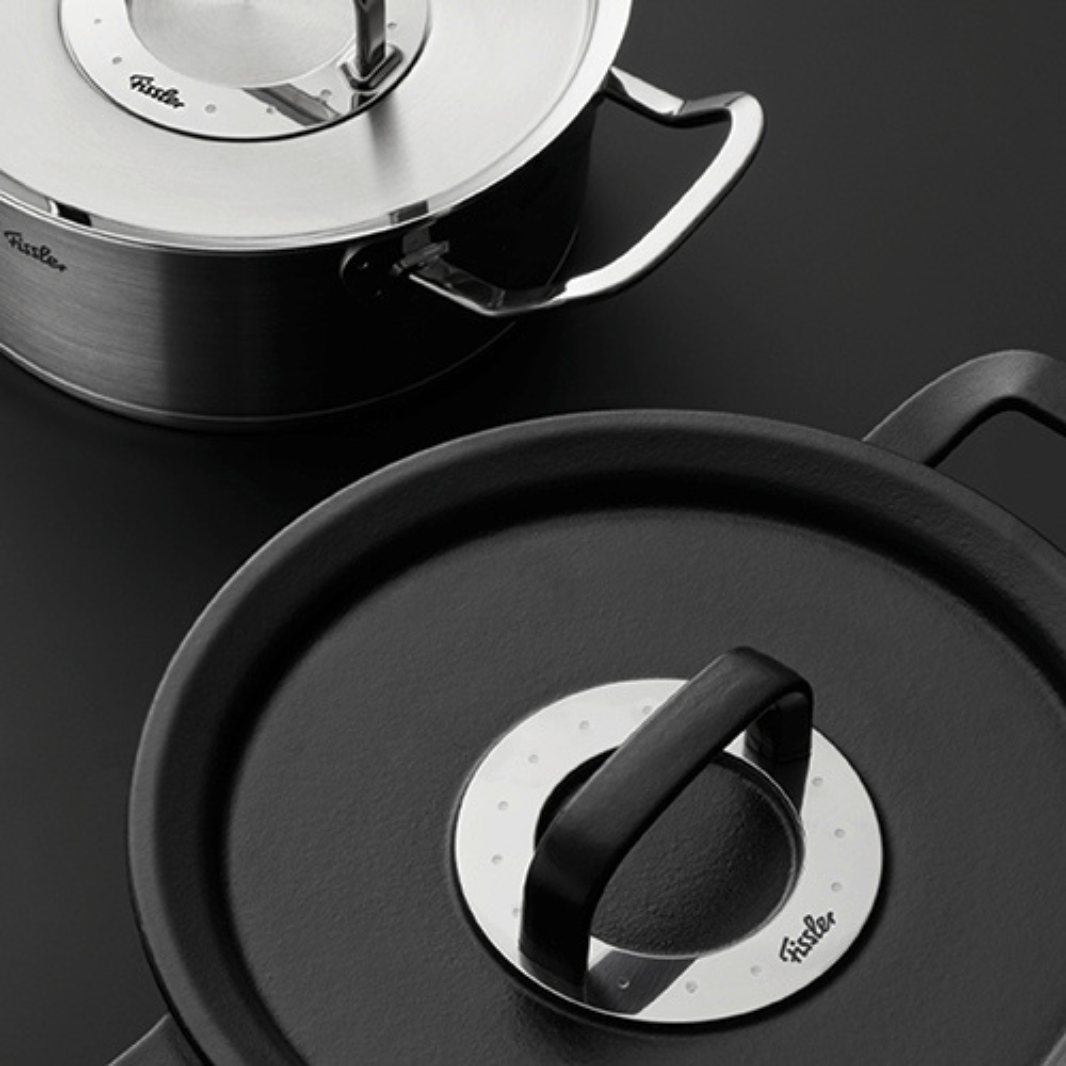 Two black Fissler pots with metal handles and branded lids, showcasing German precision and timeless design on a dark surface.