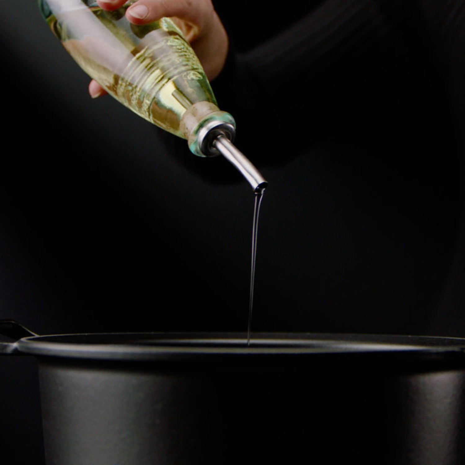 A hand pours oil from a glass bottle into a high-quality black Fissler pot, showcasing timeless German design and precise craftsmanship.