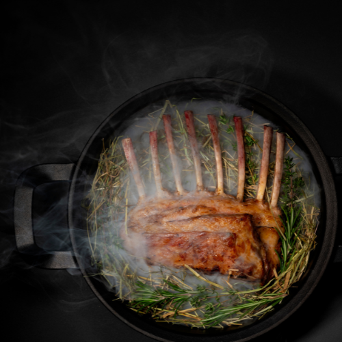 Juicy lamb on fresh herbs in a black Fissler frying pan, precisely cooked in swirling steam, against a dark background.