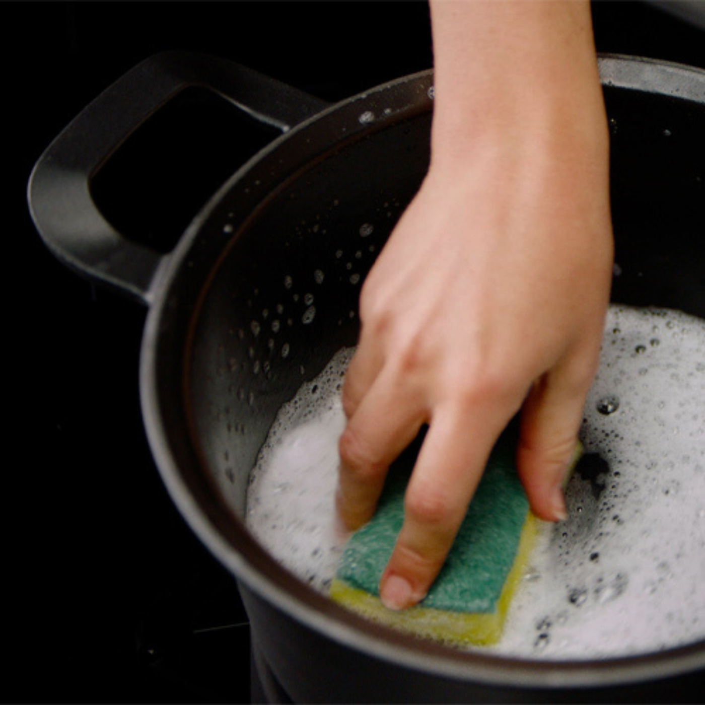 A hand cleans a high-quality black Fissler pot with a green and yellow sponge, highlighting the durability and precision of German manufacturing.