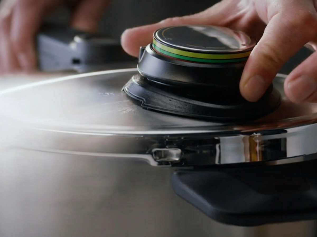 A close-up of a hand precisely adjusting the pressure valve on the lid of a Fissler stainless steel pressure cooker, showcasing German engineering and timeless design.