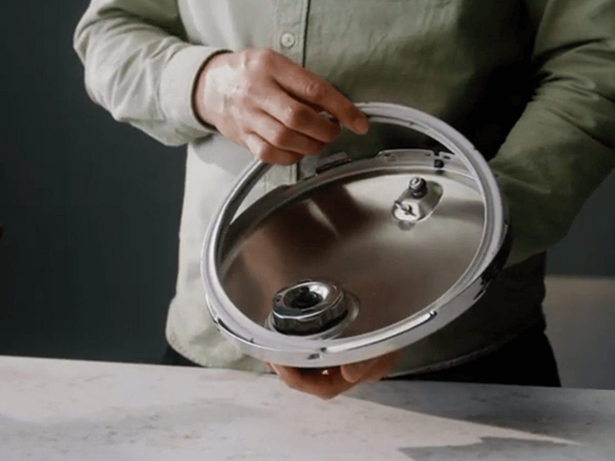 A person in a light green shirt shows a stainless steel Fissler pressure cooker lid, showcasing German engineering on a light countertop.