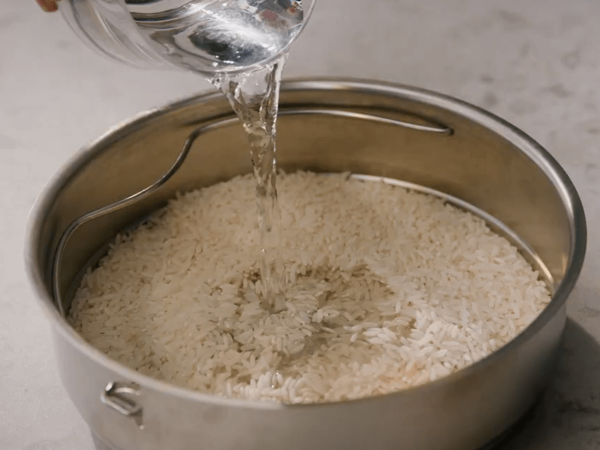 A hand pours water from a glass jug into a high-quality Fissler stainless steel pot with uncooked white rice, ready to cook.