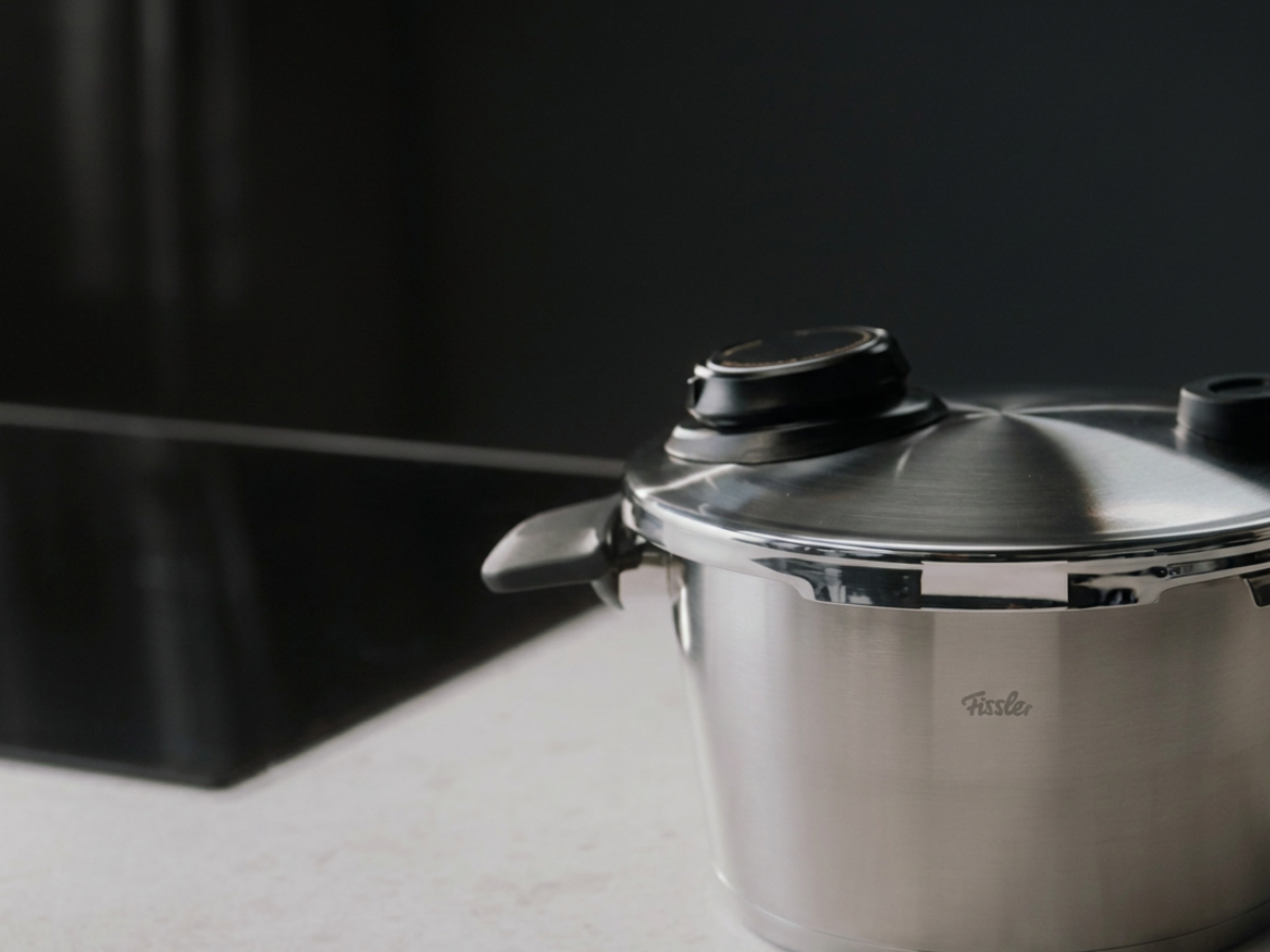 A Fissler stainless steel pressure cooker with a precision-engineered black handle rests elegantly on a light countertop beside a sleek black stovetop, set against a dark background, showcasing timeless German design and exceptional craftsmanship.