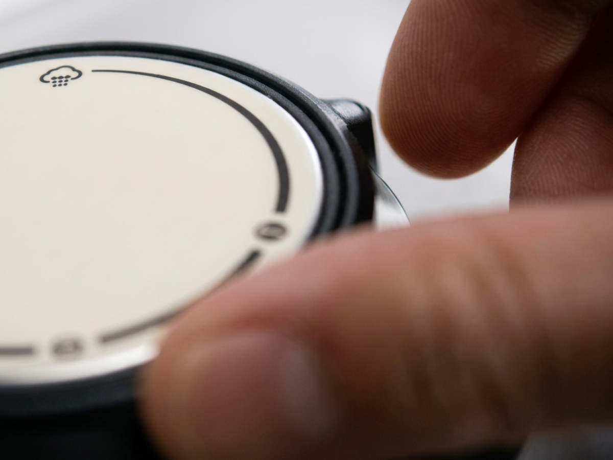 A close-up of a person’s hand holding the side of a Fissler pressure cooker lid, featuring a round, light-toned face designed with minimal markings and a small cloud icon at the top, reflecting the brand’s commitment to precision engineering and timeless German quality.