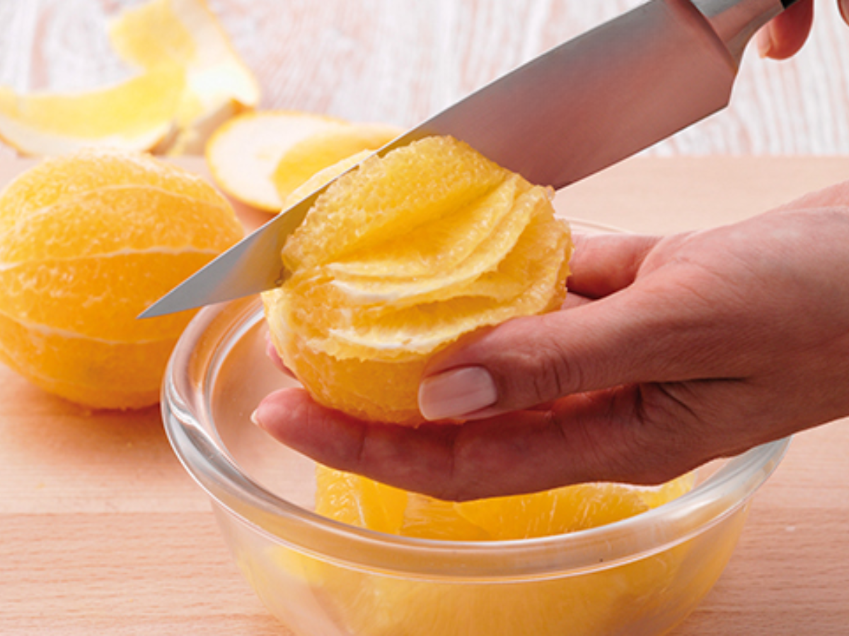 A hand precisely cuts a peeled orange over a glass Fissler bowl; orange peels lie on a wooden surface in the background.