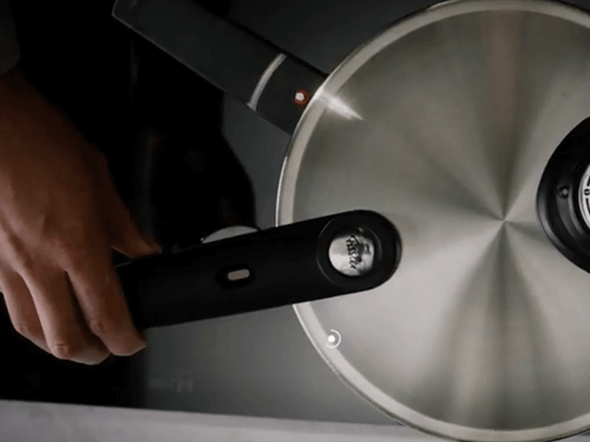 Eine Hand hält den ergonomischen schwarzen Griff eines Fissler-Edelstahltopfs mit Glasdeckel, von oben gesehen auf einem dunklen Hintergrund.