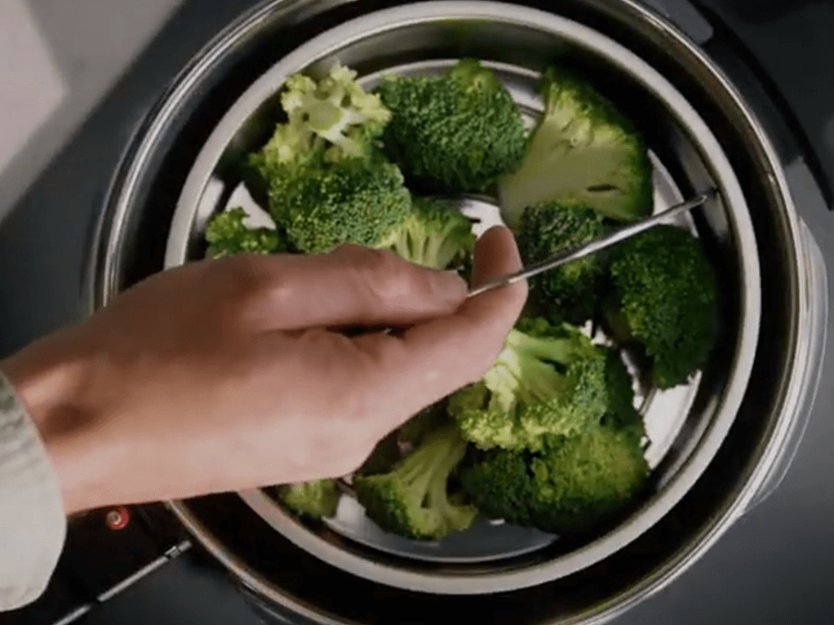 A hand lifts the lid of a Fissler stainless steel pot, revealing bright green broccoli steaming on the stove - precision and quality.