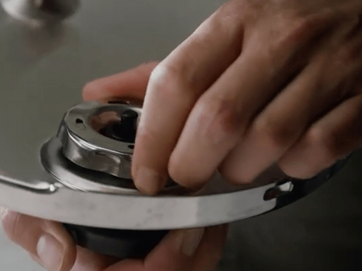 A close-up of hands precisely adjusting the stainless steel pressure regulator valve on a Fissler pressure cooker lid, showcasing German engineering and timeless design.