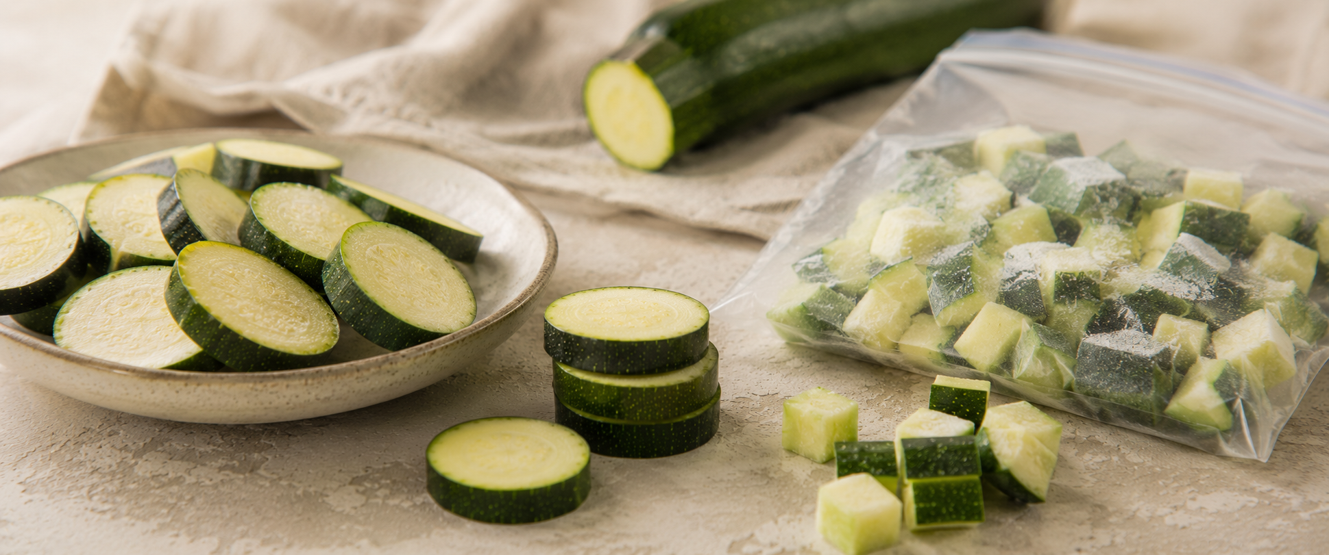Frische Zucchini, in Scheiben geschnitten und auf einem Teller angerichtet, daneben ordentlich gestapelte Zucchini-Scheiben, im Hintergrund eine ganze Zucchini und fachmännisch gewürfelte Stücke in einem durchsichtigen Beutel – all dies unterstreicht die Präzision beim Schneiden mit den hochwertigen, in Deutschland entwickelten Küchenhelfern von Fissler.