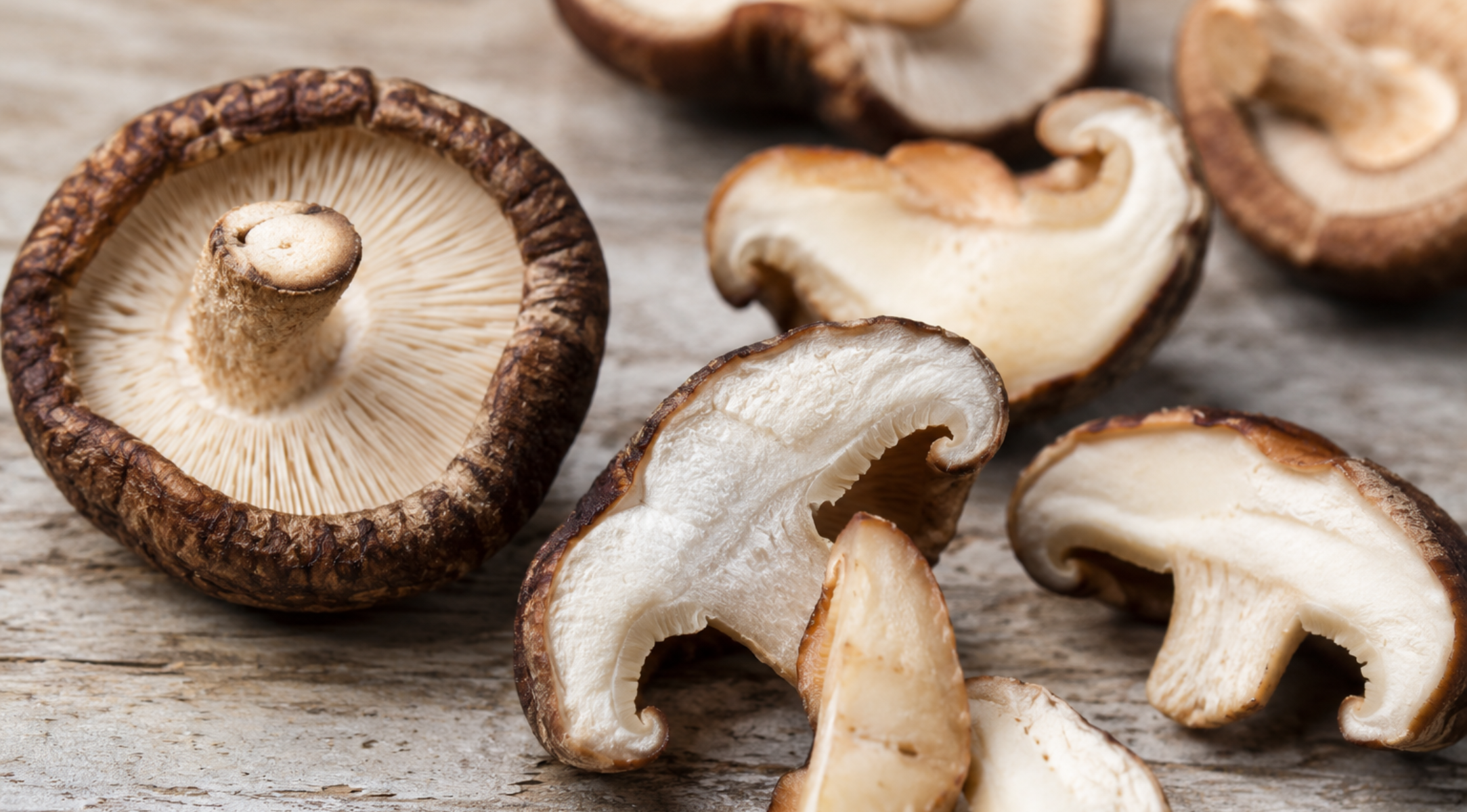 Ganze und in Scheiben geschnittene Shiitake-Pilze liegen auf einer rustikalen Holzunterlage, wodurch ihre strukturierten braunen Hüte und cremeweißen Lamellen besonders gut zur Geltung kommen – eine ideale Zutat für präzises Kochen in Fisslers hochwertigem Edelstahl-Kochgeschirr, das mit deutscher Ingenieurskunst für optimalen Geschmack und Langlebigkeit gefertigt wurde.