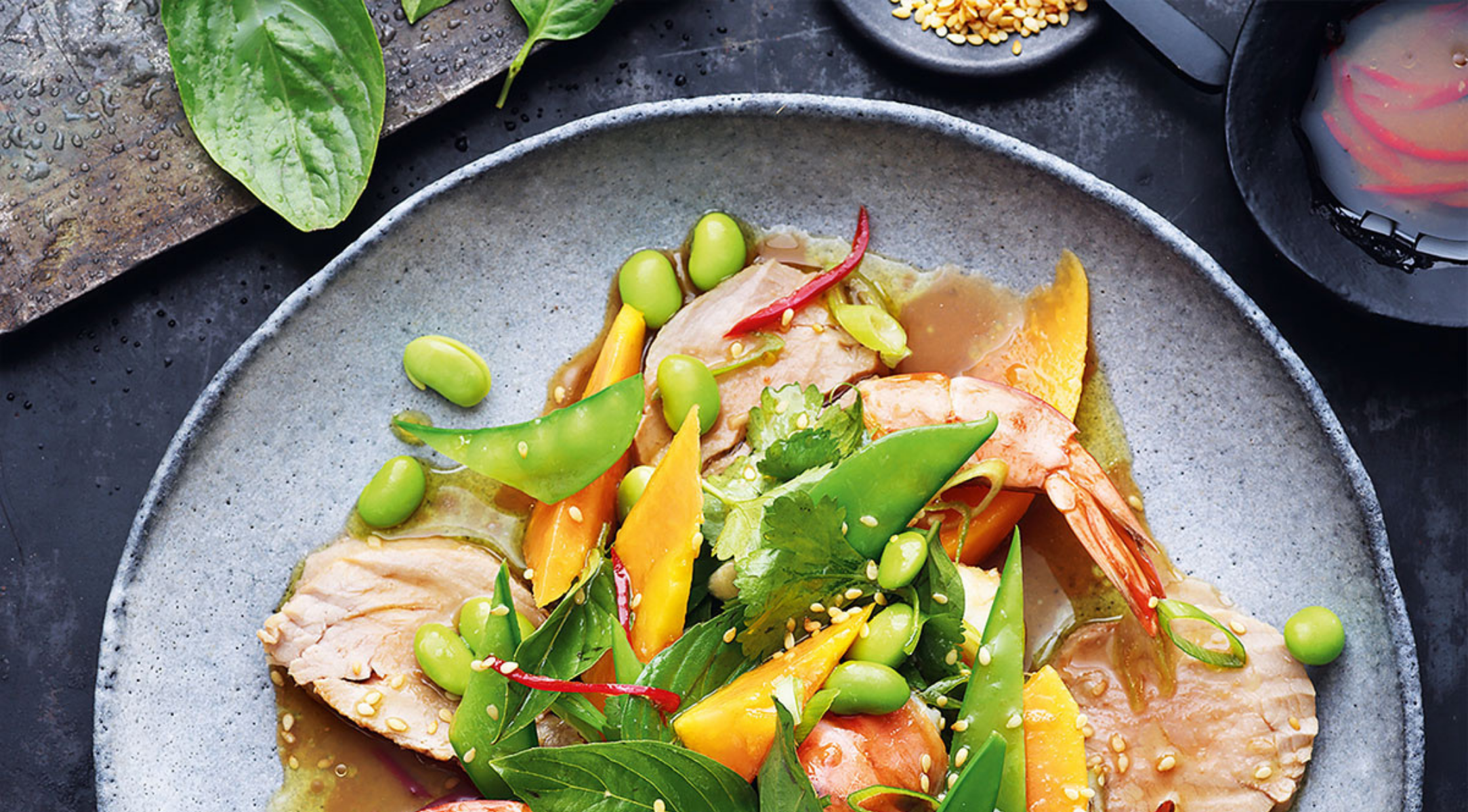 Ein lebendiger Salat mit Garnelen, Schweinefleisch, Mango und Gemüse, schön angerichtet in Fissler-Kochgeschirr, mit Soßen und Kernen an der Seite.