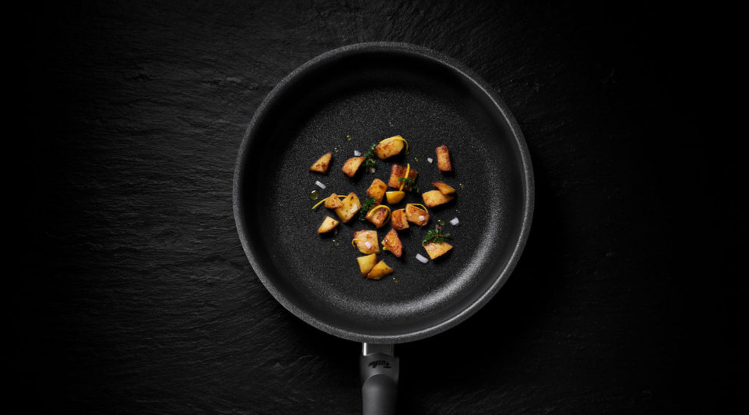 In der Mitte einer schwarzen Fissler-Pfanne liegen knusprig gebräunte Kartoffeln, ein Beispiel für deutsche Präzision vor dunkler Kulisse.