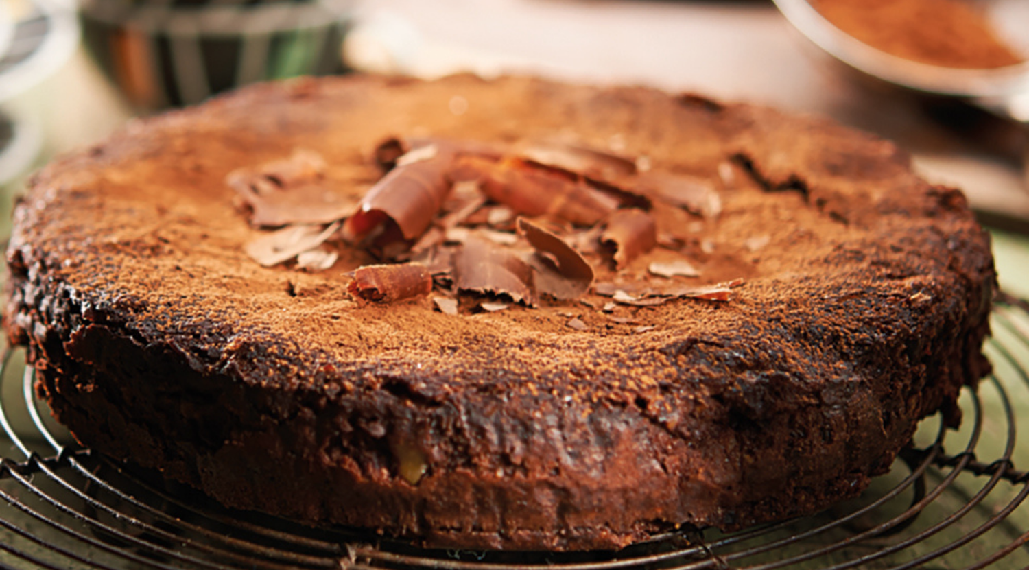 Ein runder, rustikaler Schokoladenkuchen, bestäubt mit Kakaopulver und Schokoladenspänen, kühlt auf einem Fissler-Drahtgitter ab und zeigt deutsche Qualität.