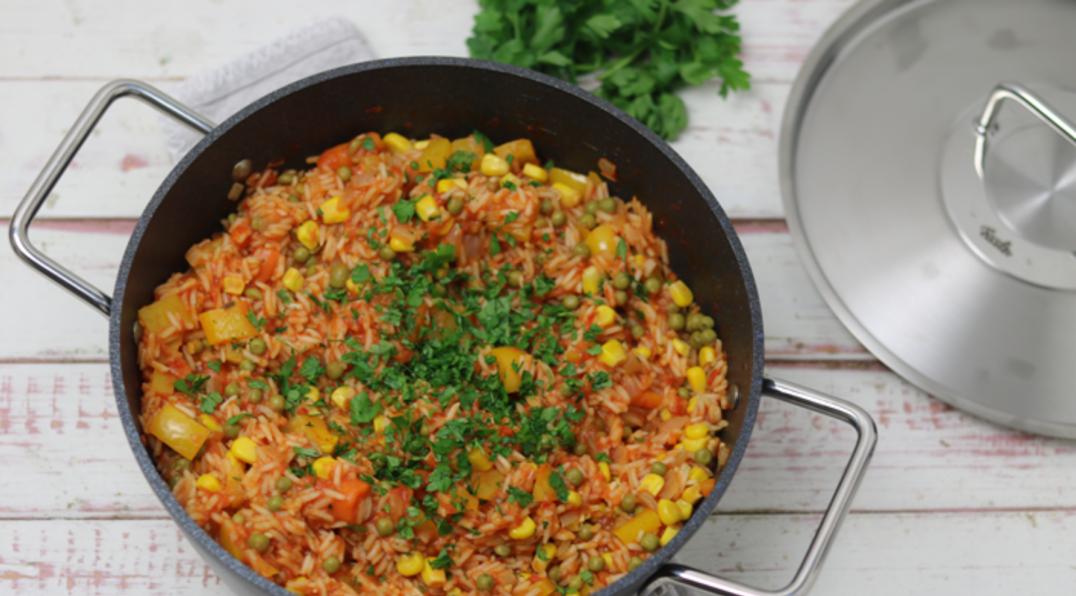Schwarzer Fissler-Topf mit Tomatenreis, Mais und Paprika auf weißem Holz, daneben frische Petersilie und ein typischer Fissler-Deckel.