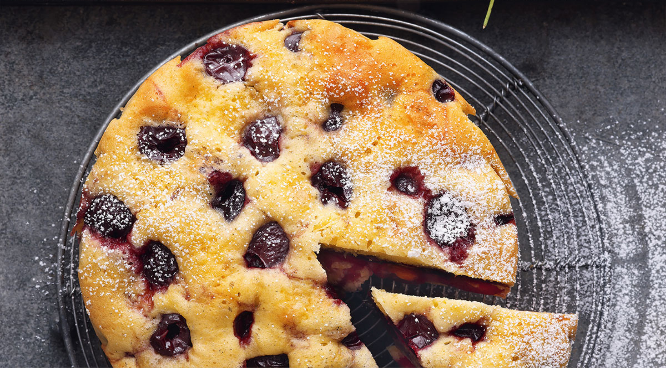 Ein runder Kirschkuchen mit goldener Kruste, mit Zucker bestäubt, eine Scheibe auf einem Fissler-Edelstahl-Kühlrost herausgezogen.