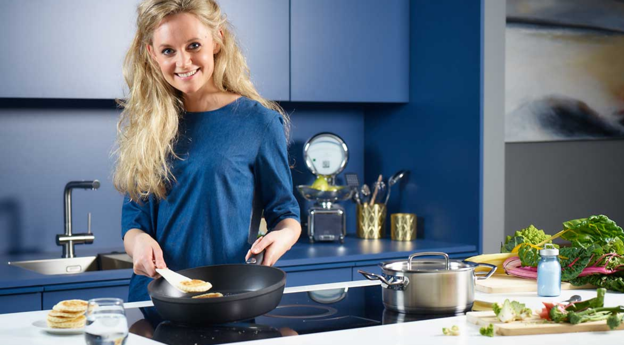 Eine Frau mit langen blonden Haaren backt Pfannkuchen in einer hochwertigen Fissler-Pfanne in einer schlichten blauen Küche, Fissler-Topf und -Geräte in der Nähe.