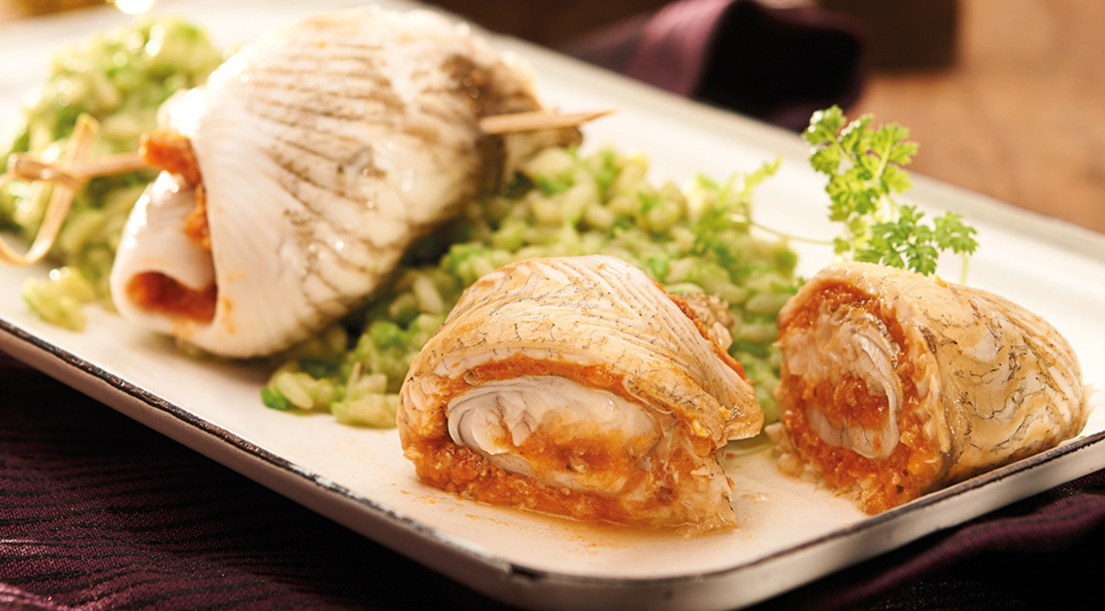 Zwei gerollte weiße Fischfilets mit Tomatenfüllung auf einem Tablett, serviert mit Grünzeug, präsentiert mit der Präzision und dem zeitlosen Stil von Fissler.