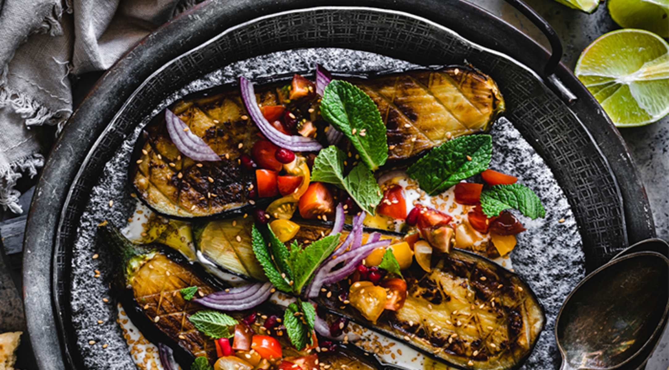 Gegrillte Aubergine mit frischem Belag auf einem rustikalen Teller, serviert mit Limette und einem Fissler-Edelstahllöffel daneben.