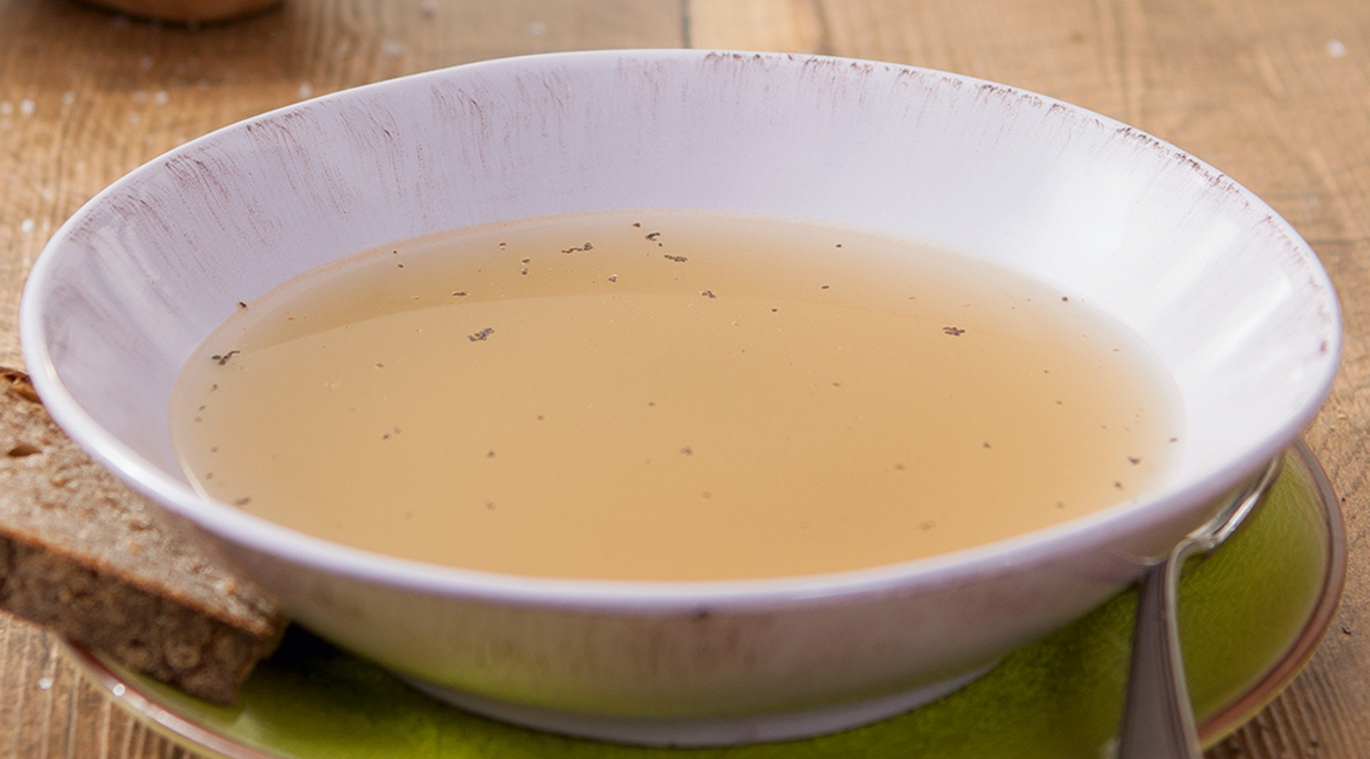Eine flache weiße Schale mit Kräuterbrühe steht auf einem grünen Teller mit Brot und einem Löffel, der auf einem Holztisch in Fissler-Qualität steht.