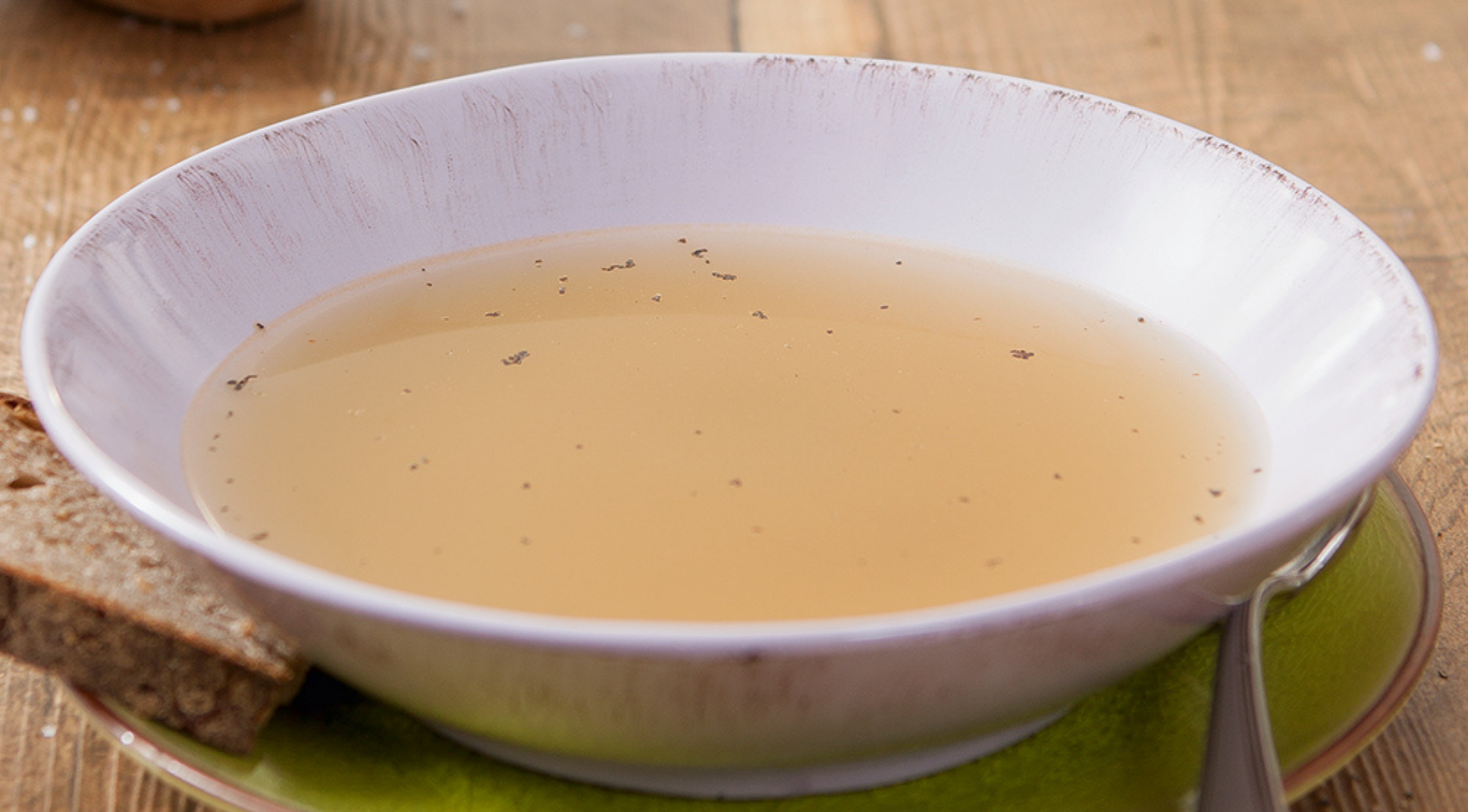Eine flache weiße Schale mit Kräuterbrühe steht auf einem grünen Teller mit Brot und einem Löffel, der auf einem Holztisch in Fissler-Qualität steht.