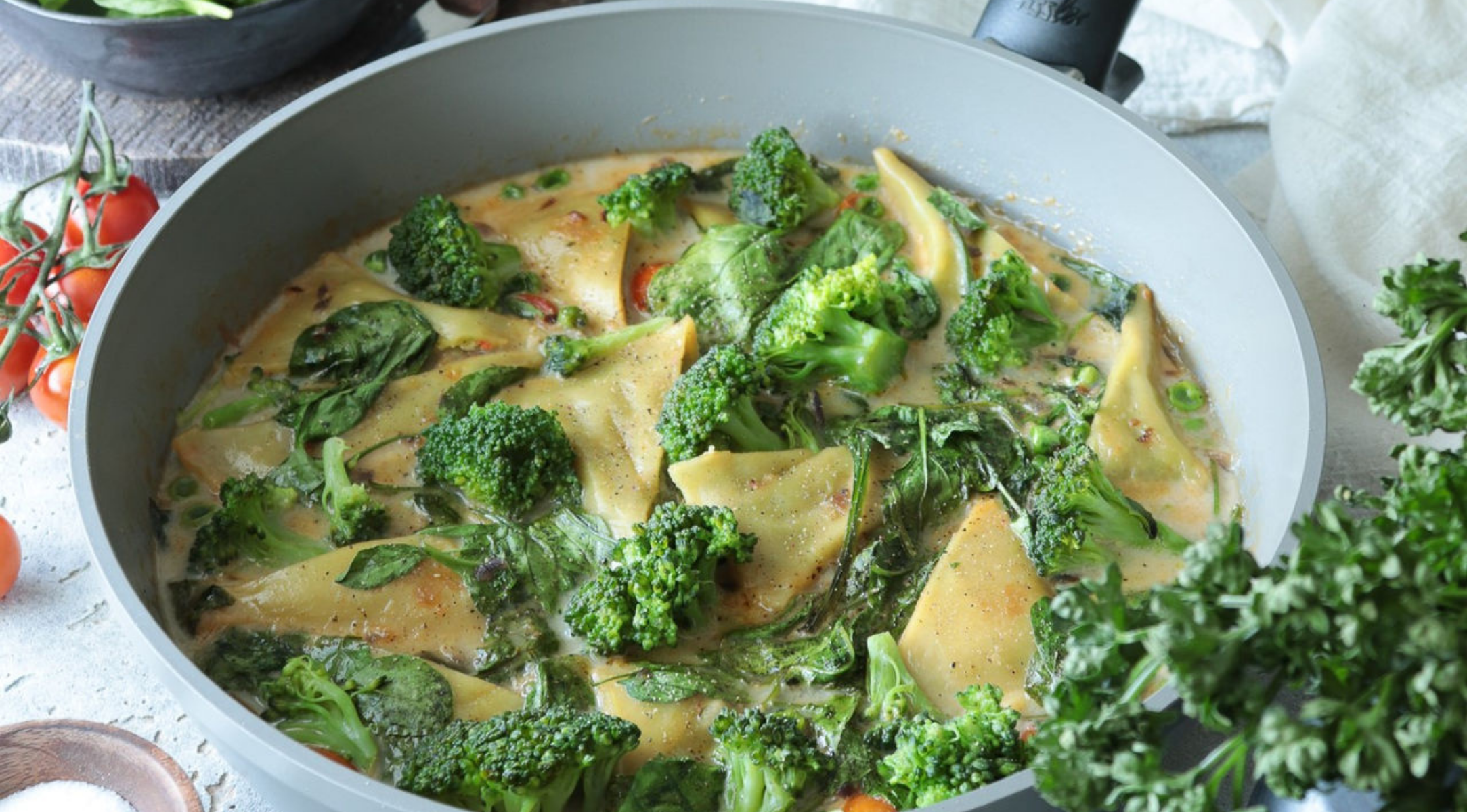 Eine Fissler-Pfanne mit cremigen Nudeln, Brokkoli, Spinat und Soße; frisches Gemüse und Kräuter umgeben unser deutsches Präzisionskochgeschirr.