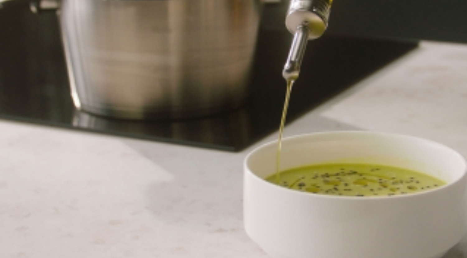 In einer weißen Schale träufelt Olivenöl in die grüne Suppe, dahinter steht ein mit deutscher Präzision gefertigter Fissler-Edelstahltopf.