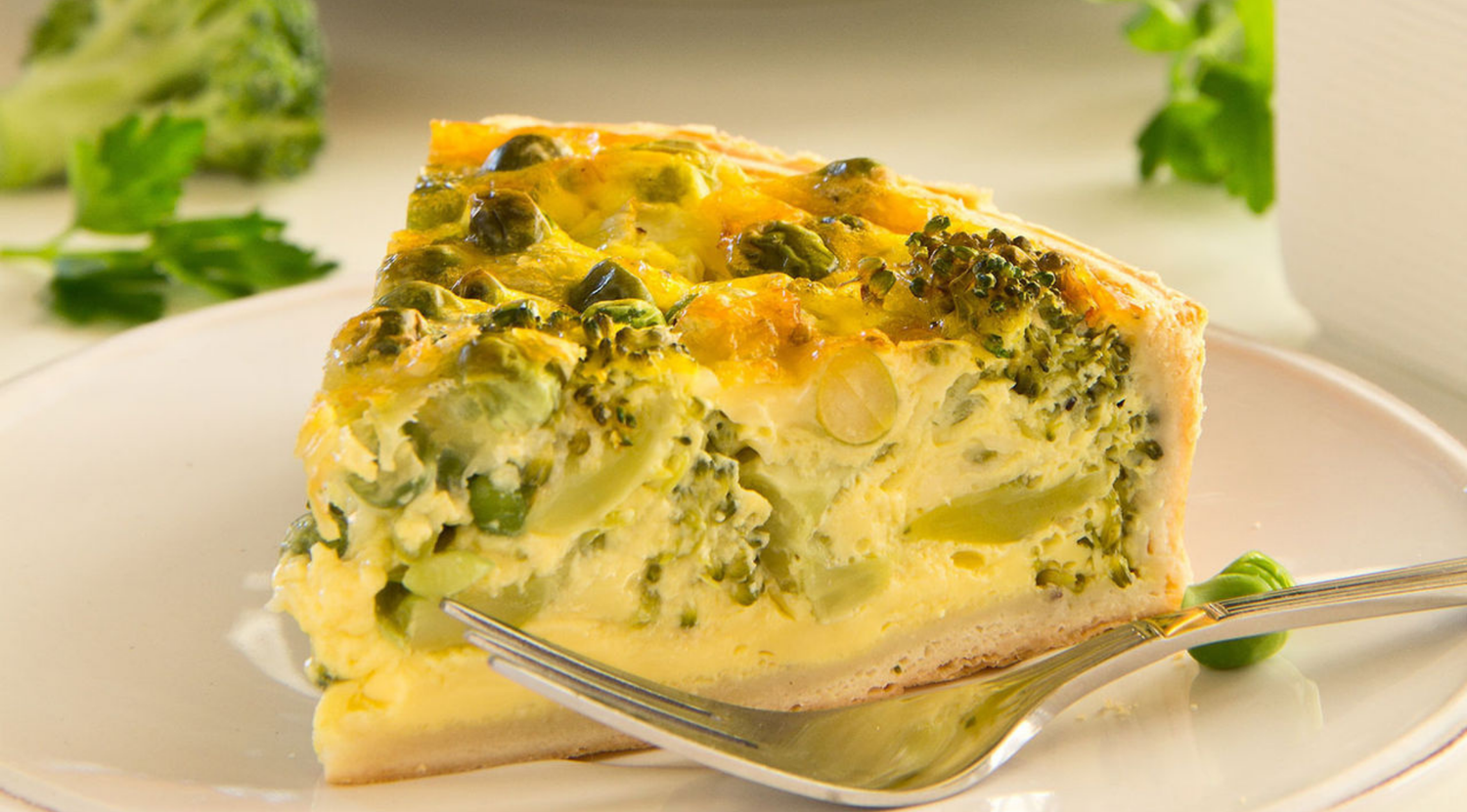 Ein Stück goldene Brokkoli-Quiche auf einem weißen Teller, garniert mit Kräutern, perfekt gebacken im Fissler-Qualitätskochgeschirr.
