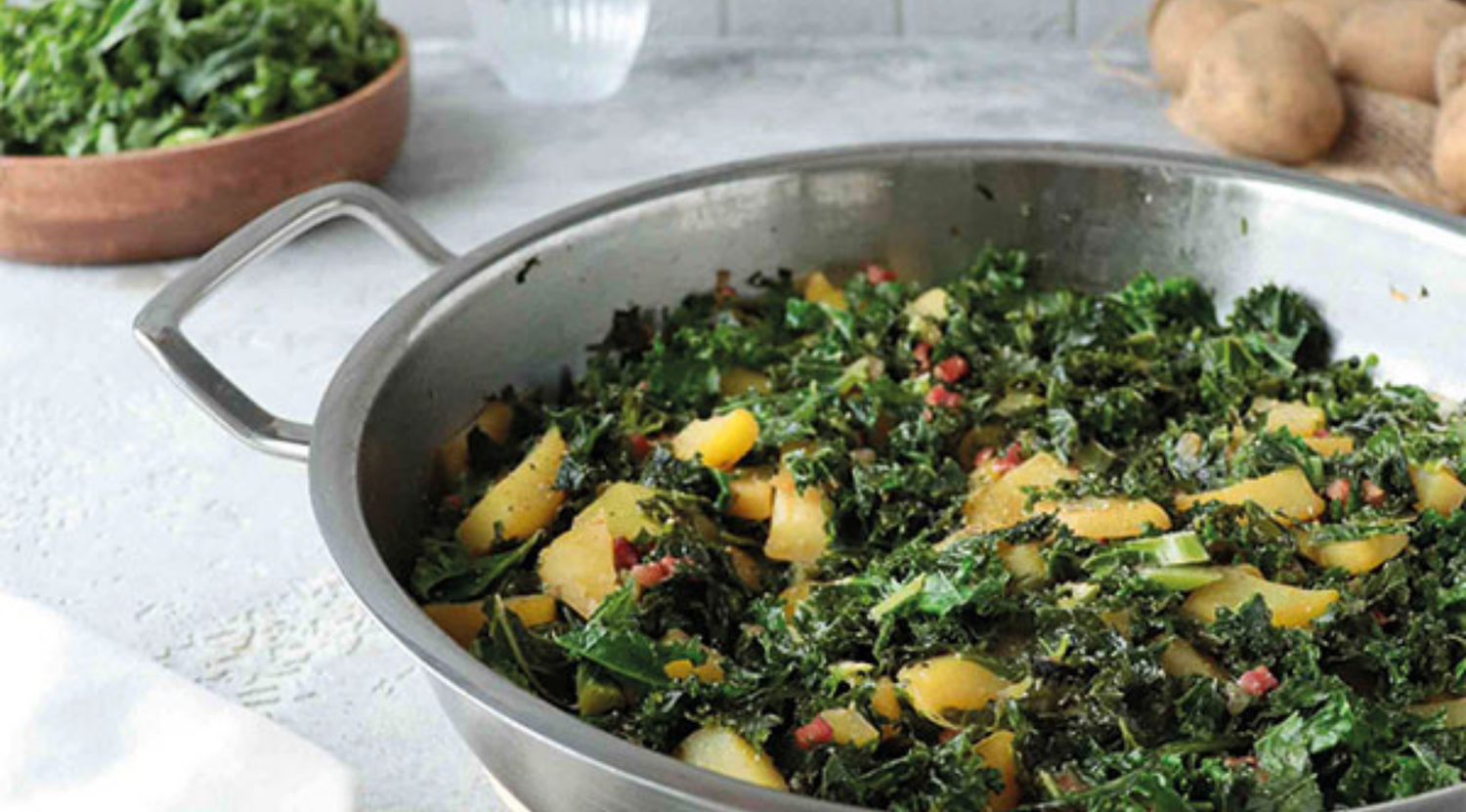 Eine Fissler-Edelstahlpfanne mit gebratenem Grünkohl, Kartoffeln und Speck auf einer Theke; im Hintergrund frisches Grün und Kartoffeln.