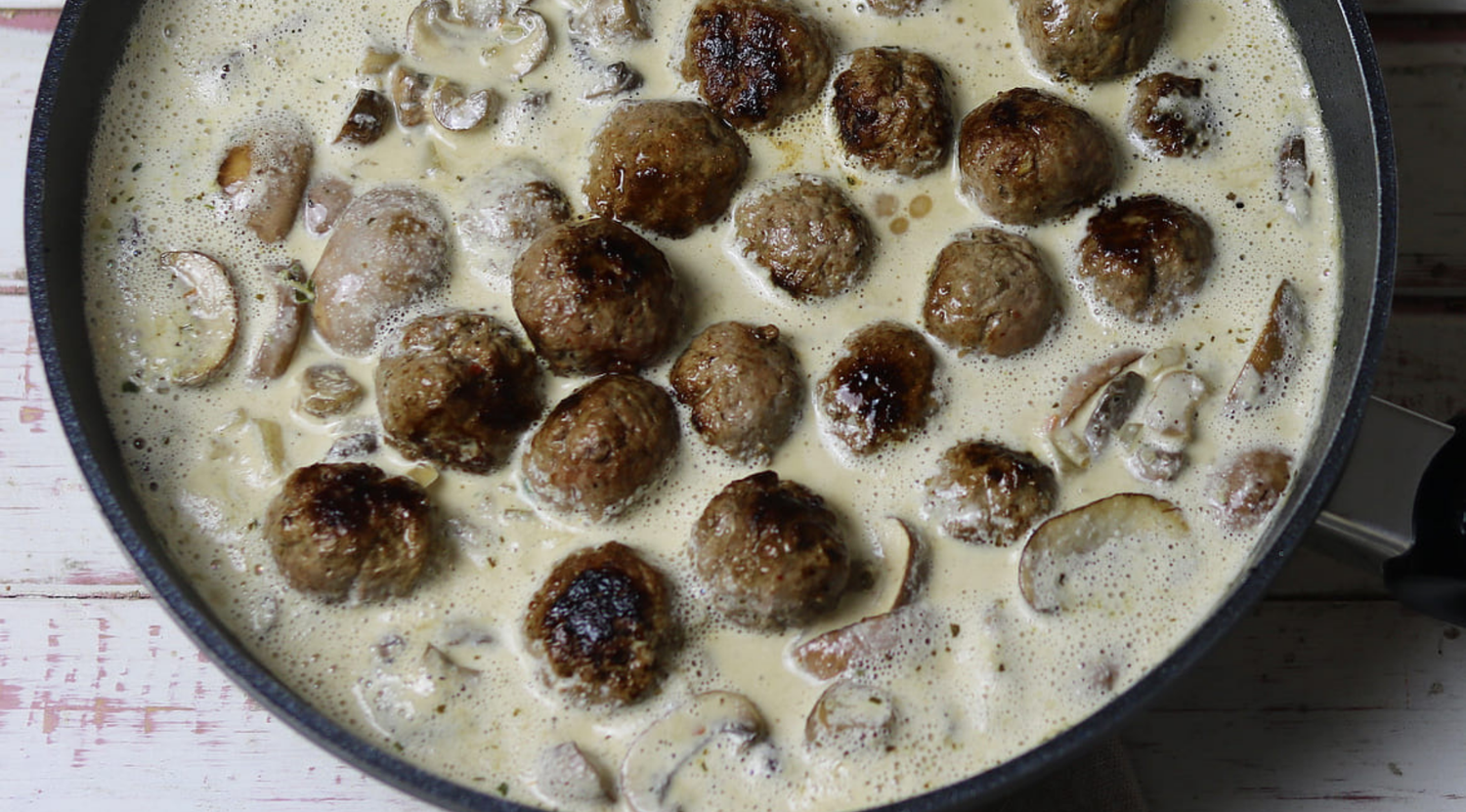 Eine hochwertige Fissler-Pfanne mit cremiger Sauce, köchelnden Fleischbällchen und Pilzen auf einer hellen Holzoberfläche - deutsches Präzisionskochgeschirr.