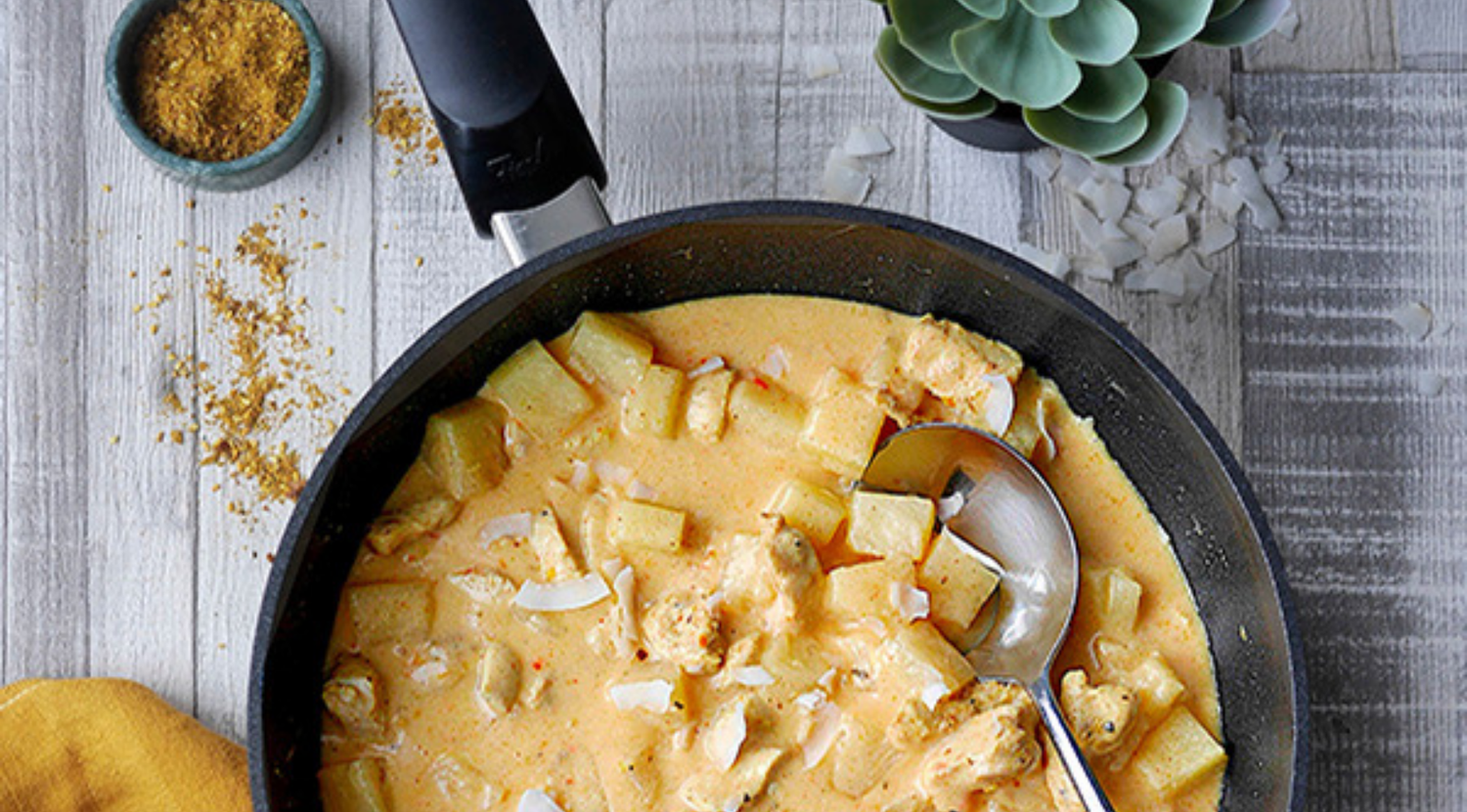 Eine hochwertige Fissler-Pfanne mit cremigem Hühner- und Kartoffelcurry, umgeben von Currypulver, Kokosraspeln und einer gelben Serviette.