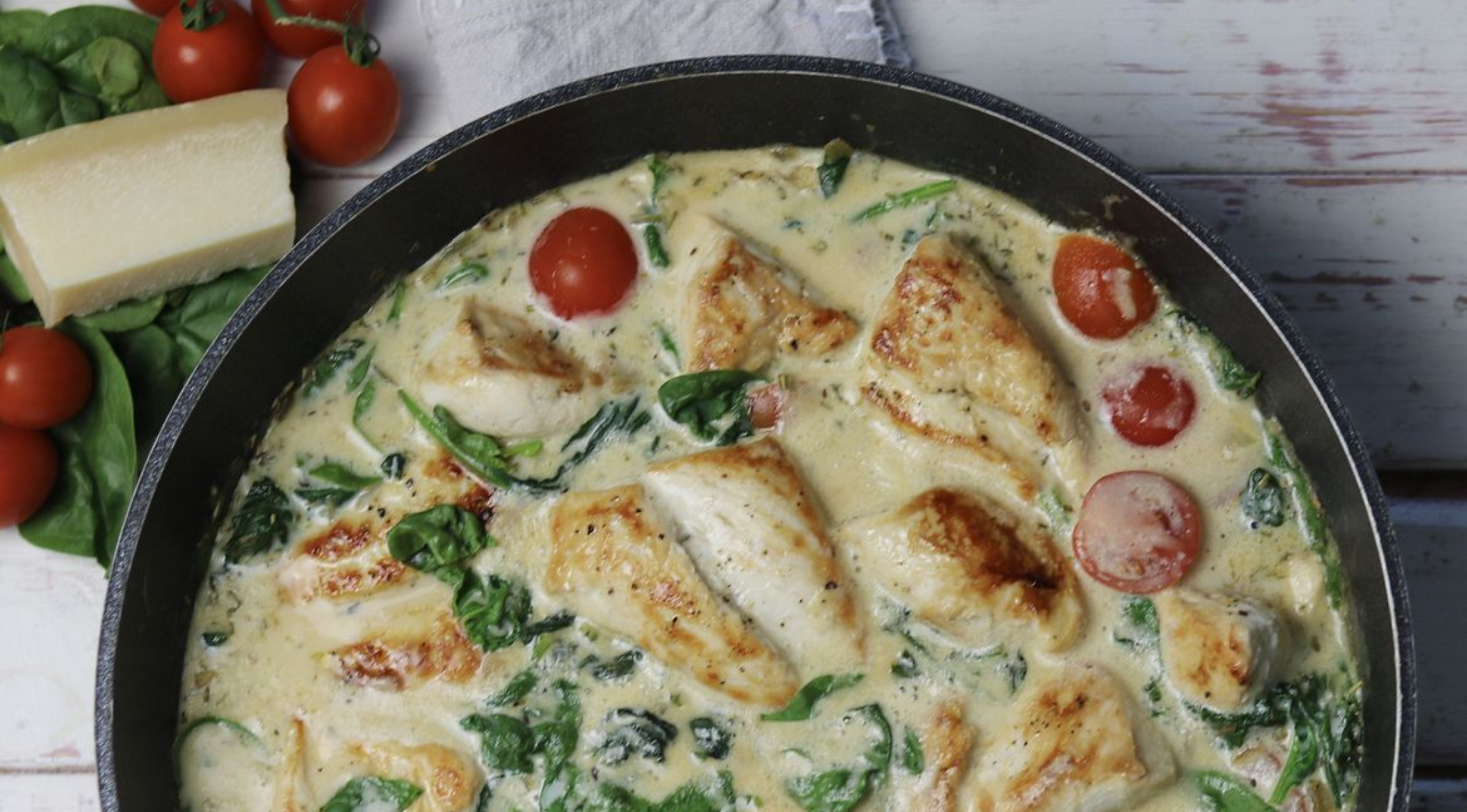 Eine Fissler-Pfanne zeigt cremiges Hähnchen, Kirschtomaten und Spinat in weißer Soße, dazu frisches Grün und Käse.
