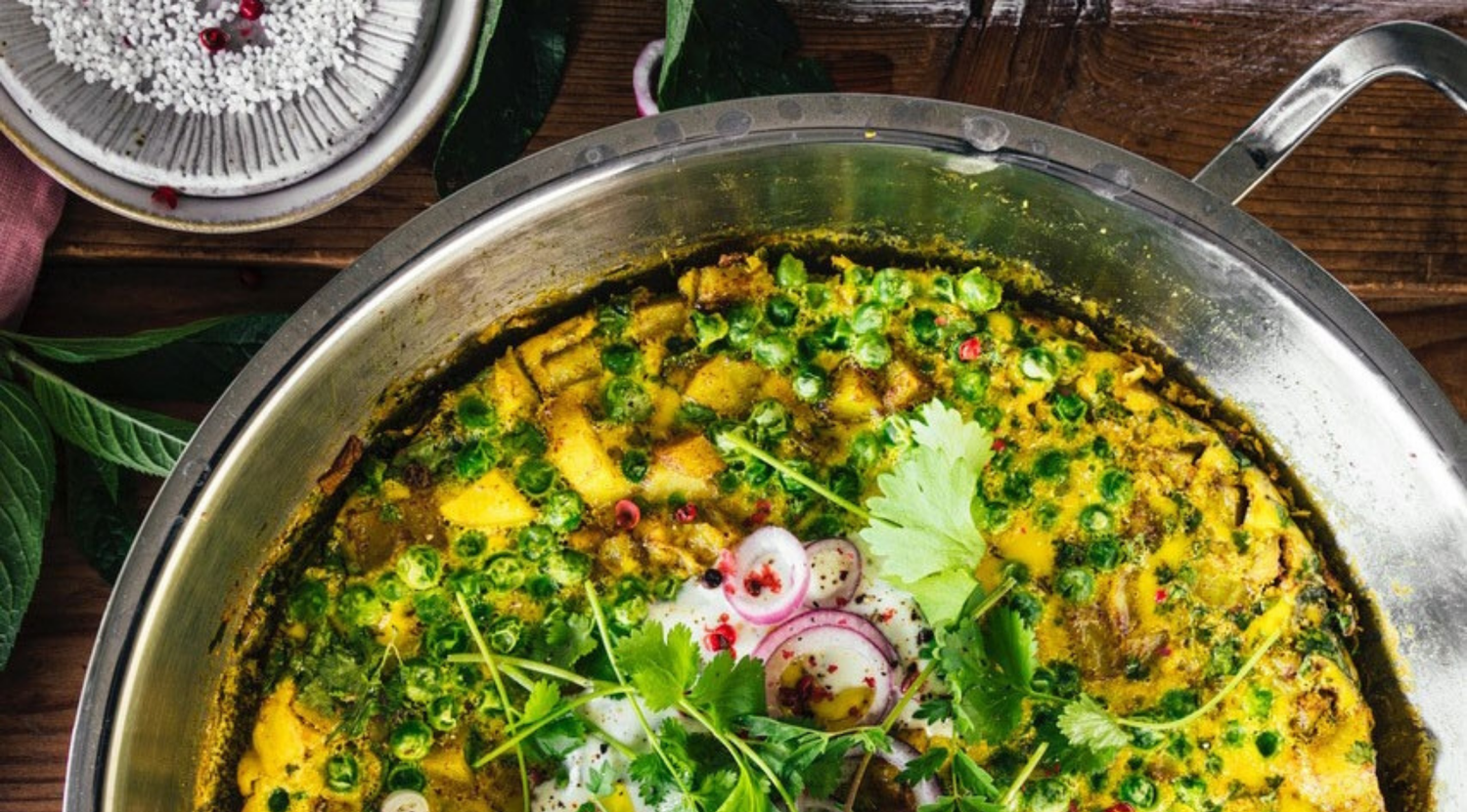 Fissler-Edelstahlpfanne mit leuchtend gelbem Curry, frischen Kräutern und Gemüse auf einem Holztisch, der deutsche Handwerkskunst demonstriert.