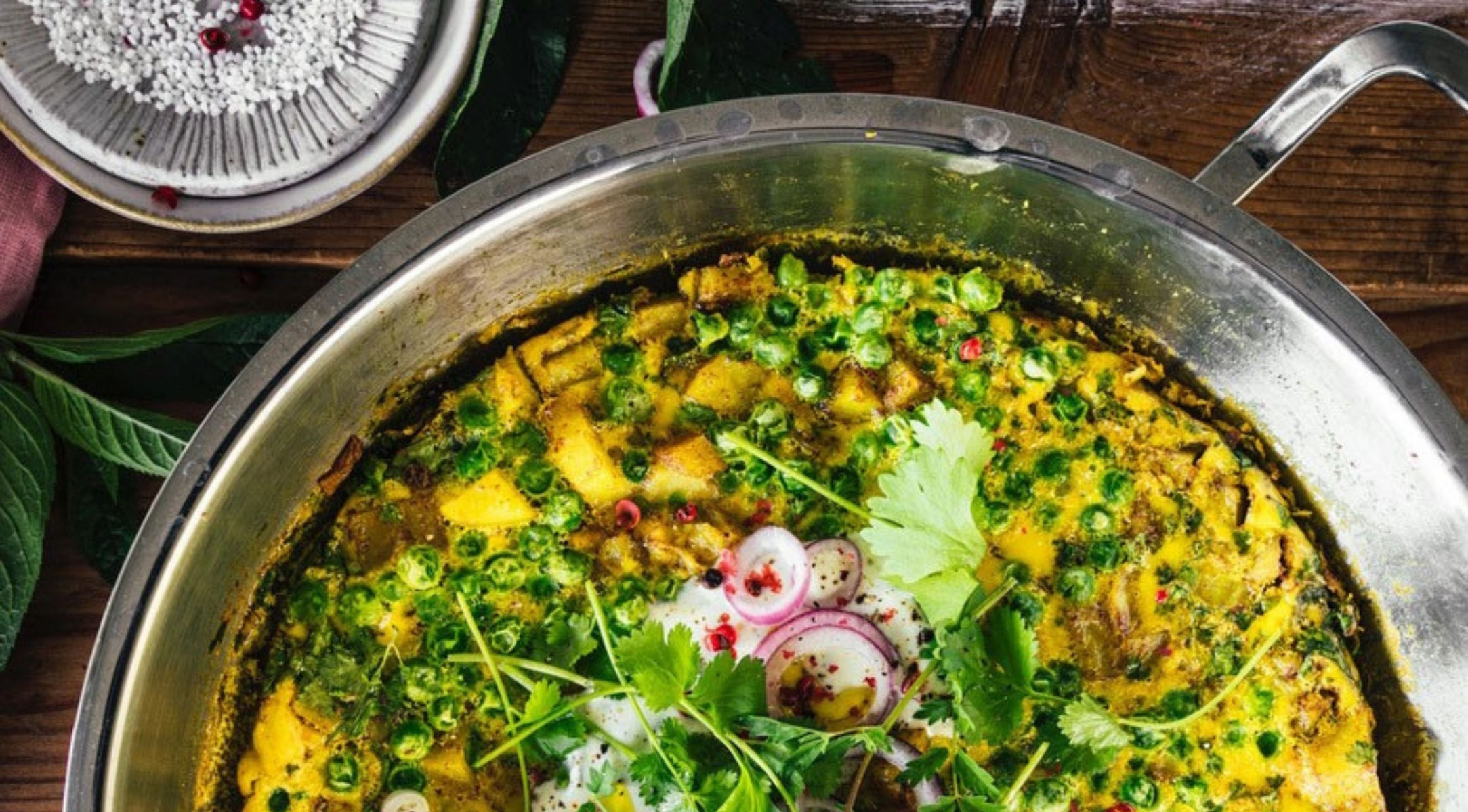 Fissler-Edelstahlpfanne mit leuchtend gelbem Curry, frischen Kräutern und Gemüse auf einem Holztisch, der deutsche Handwerkskunst demonstriert.