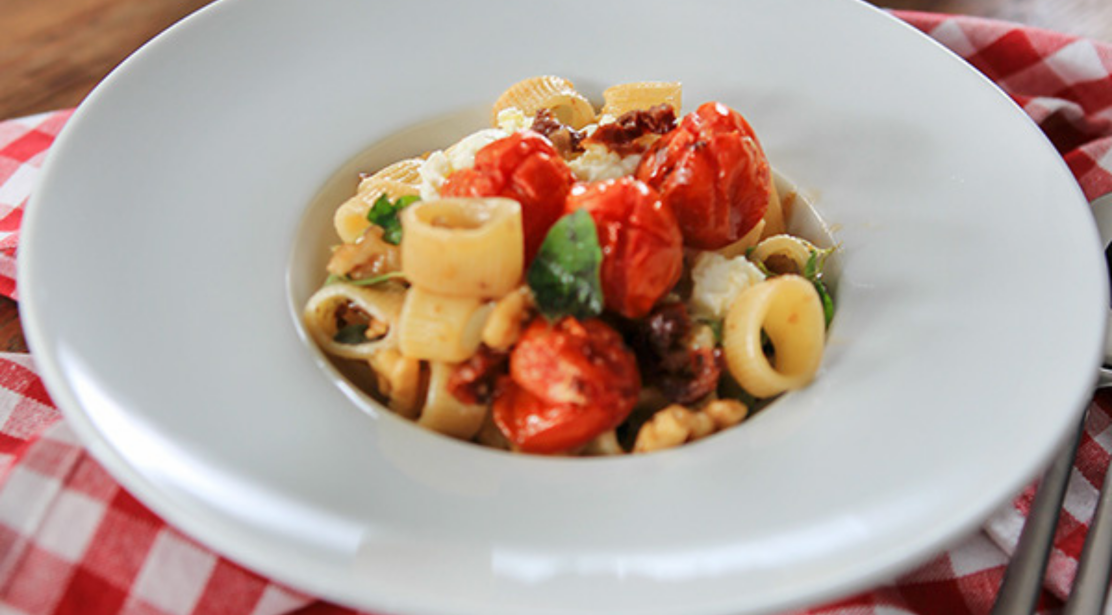 Pasta mit gerösteten Kirschtomaten, Walnüssen, Basilikum und Käse auf einem weißen Teller auf einem rot-karierten Stoff-Fissler Kochgeschirr Eleganz.