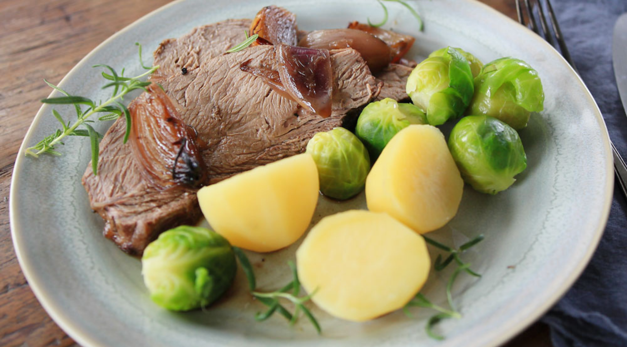 Genießen Sie Roastbeef mit Zwiebeln, Kartoffeln und Rosenkohl - zubereitet im Präzisionskochgeschirr von Fissler auf einem Holztisch.